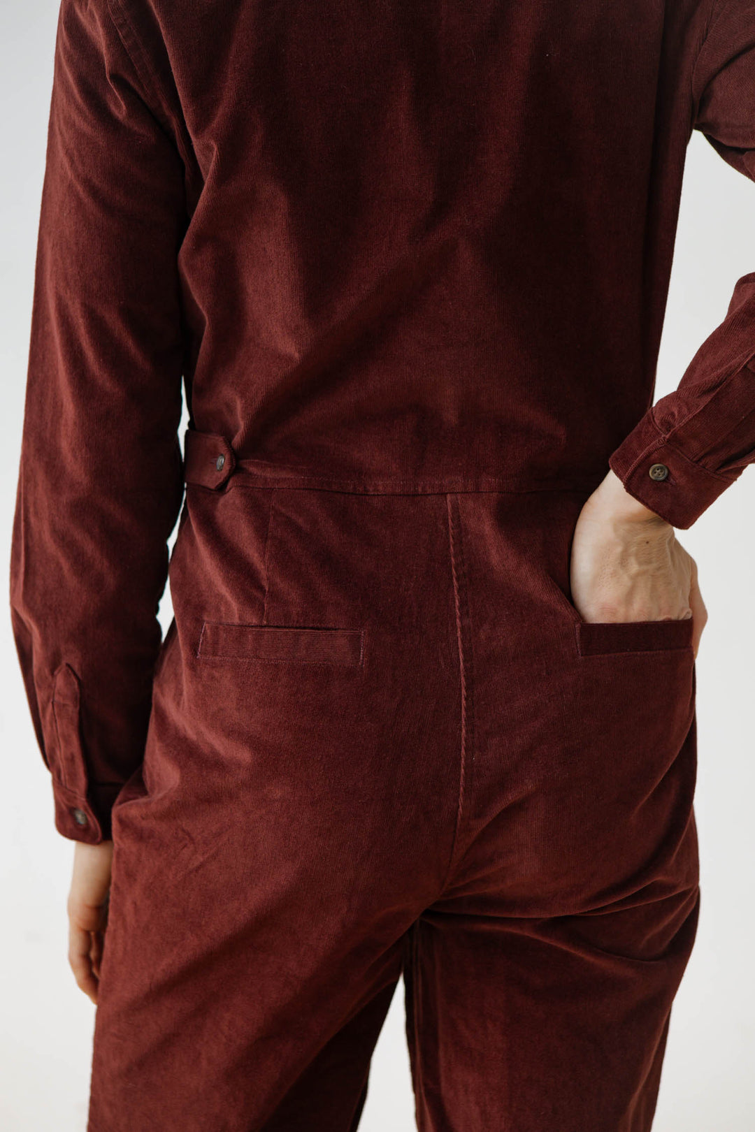 A person stands with their back to the camera, one hand in a pocket, wearing the Bridge & Burn Faye Jumpsuit in Dark Red Pinwale, effortlessly showcasing this stylish women's corduroy workwear piece.