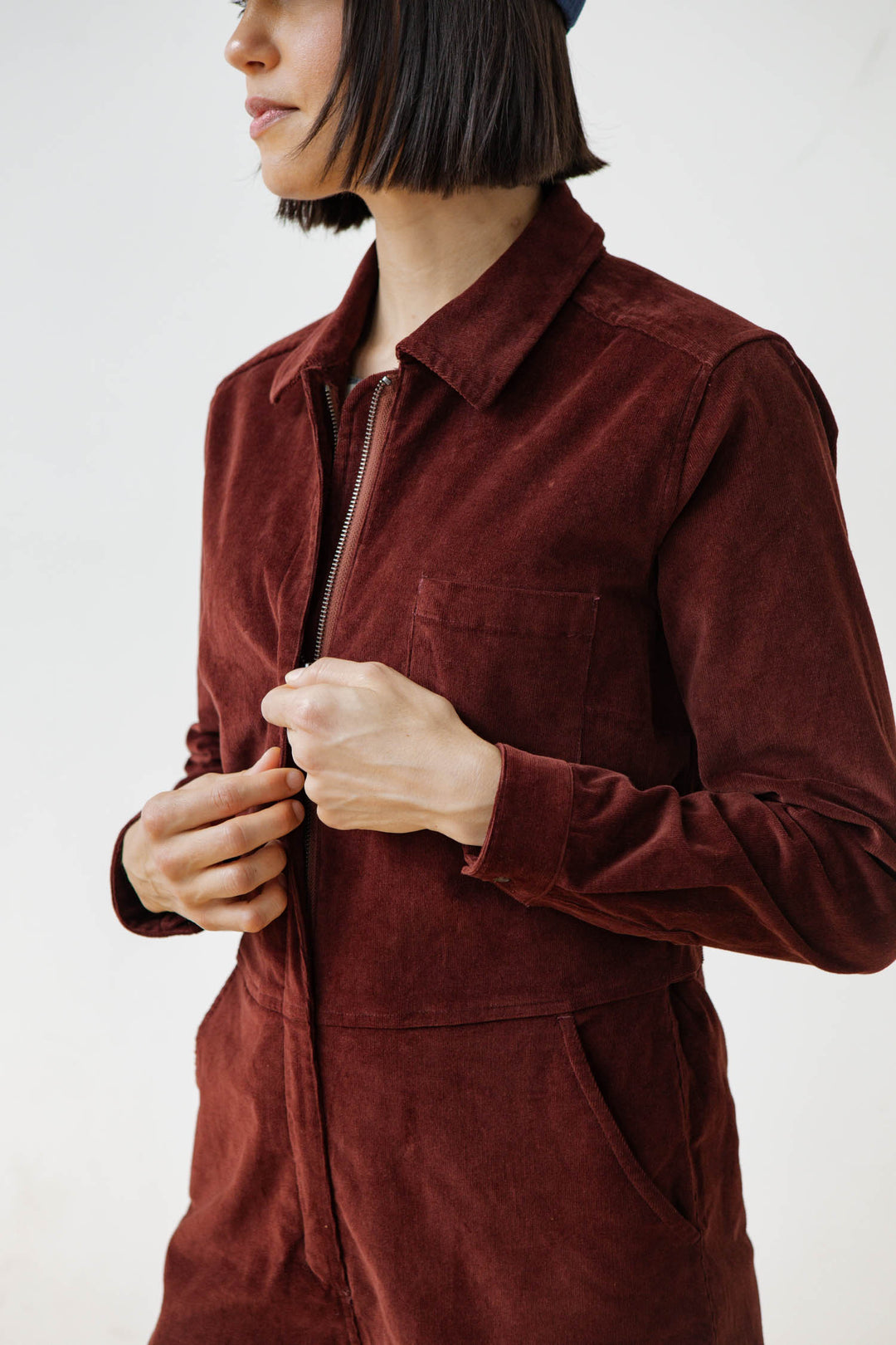 A person with straight, shoulder-length dark hair models the Bridge & Burn Faye Jumpsuit in Dark Red Pinwale, partially unzipped at the front, standing against a plain white background with their face partially out of frame.