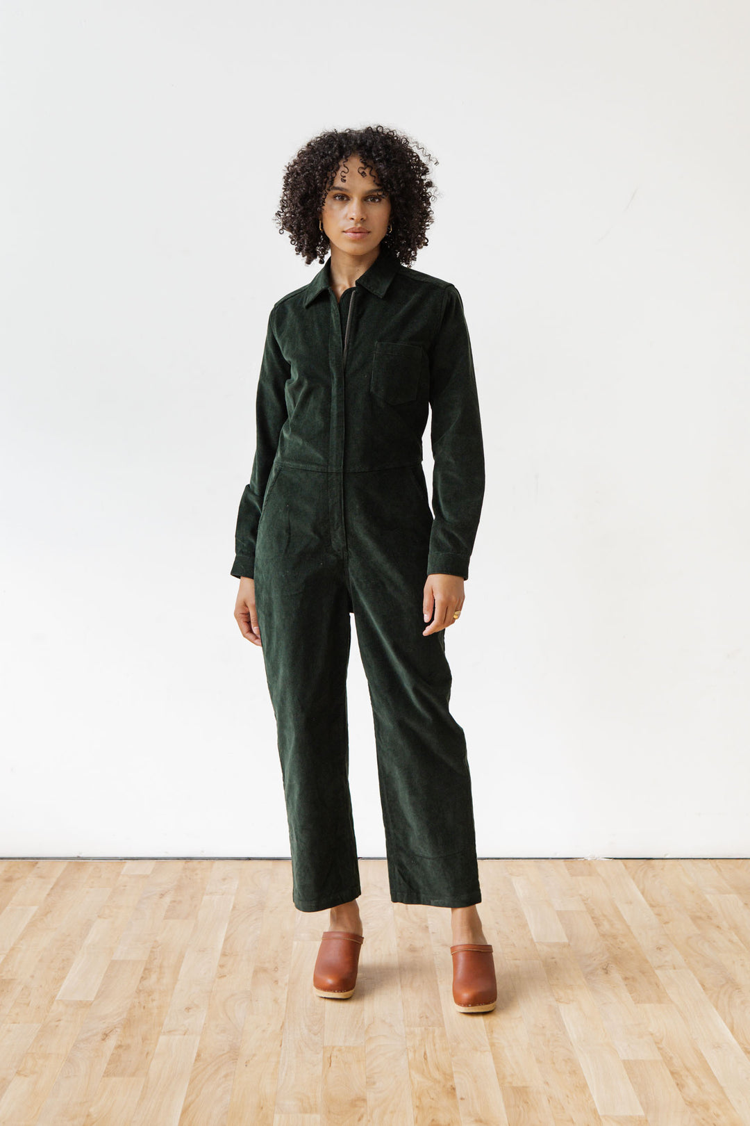 A person with curly hair stands indoors on a wooden floor, wearing the Bridge & Burn Faye Jumpsuit in Deep Pine Pinwale and brown clogs, facing the camera against a plain white wall.