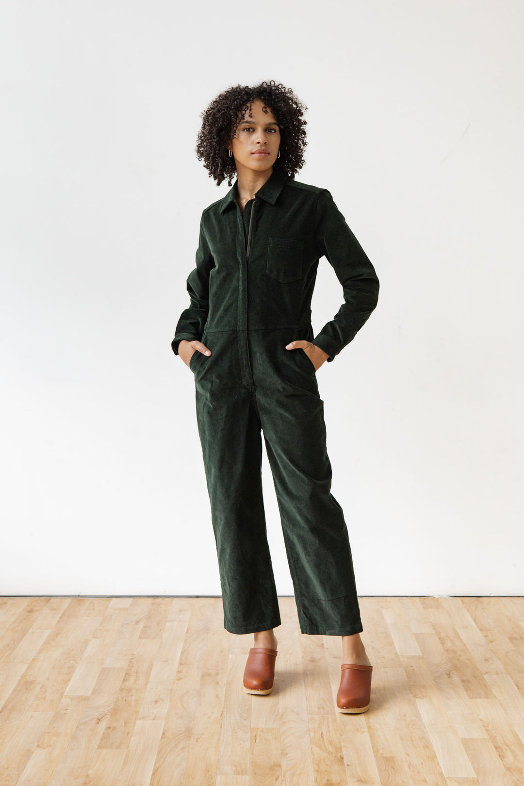 Standing on a wooden floor against a white background, a person with curly hair wears the Bridge & Burn Faye Jumpsuit in Deep Pine Pinwale and brown clogs, hands in pockets, looking to the side.