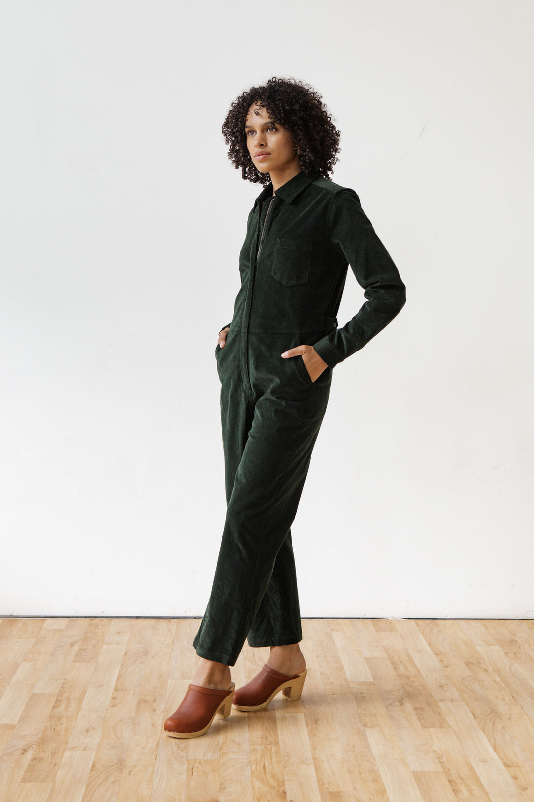 A person with curly hair wears the Bridge & Burn Faye Jumpsuit in Deep Pine Pinwale, paired with brown clogs, standing on a light wooden floor against a plain white background with hands in their pockets.