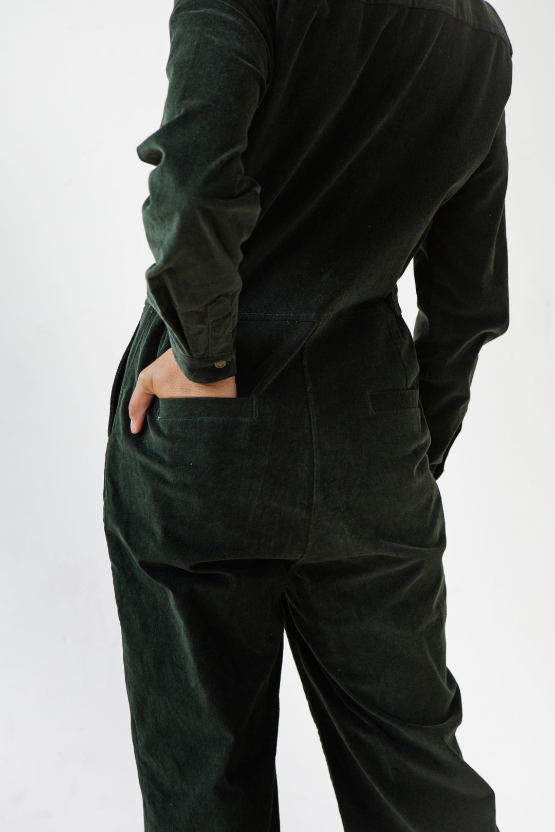 A person in the Bridge & Burn Faye Jumpsuit in Deep Pine Pinwale stands with one hand in their back pocket, shown from behind against a plain white background.