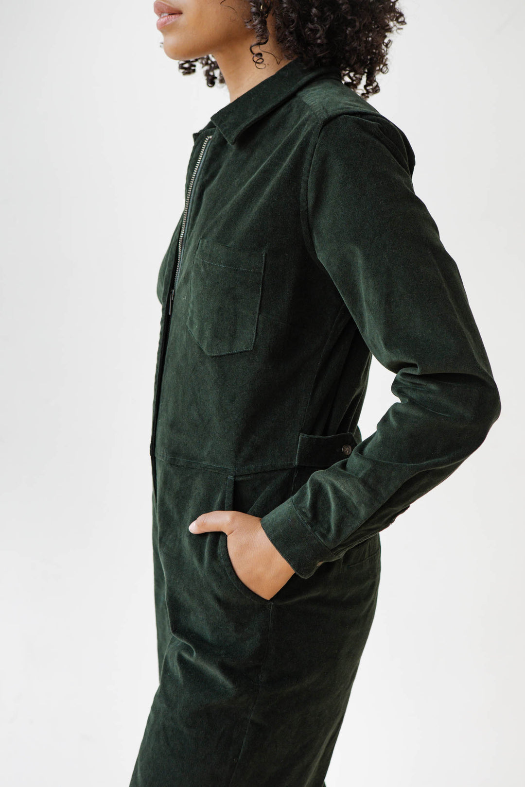 A person with curly hair wears the Bridge & Burn Faye Jumpsuit in Deep Pine Pinwale, hands in side pockets, standing against a plain white background.