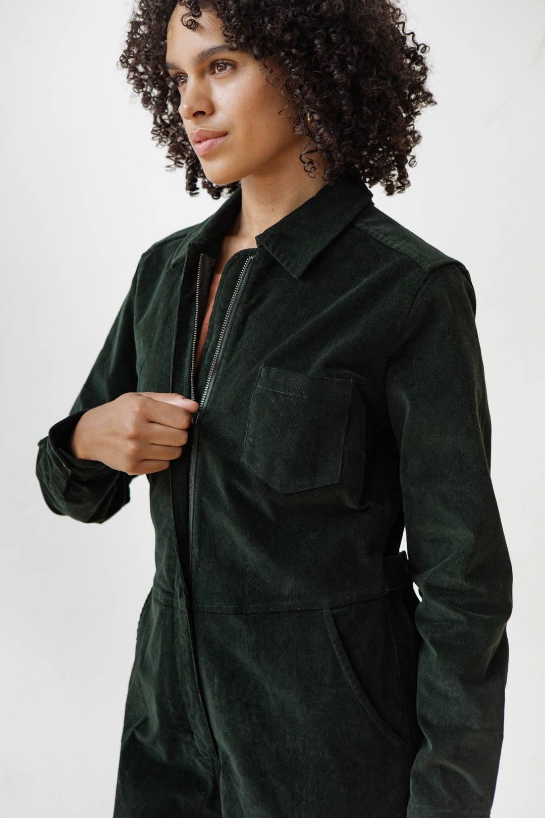A woman with curly hair wears the Bridge & Burn Faye Jumpsuit in Deep Pine Pinwale, standing against a plain white background and looking slightly to the side.