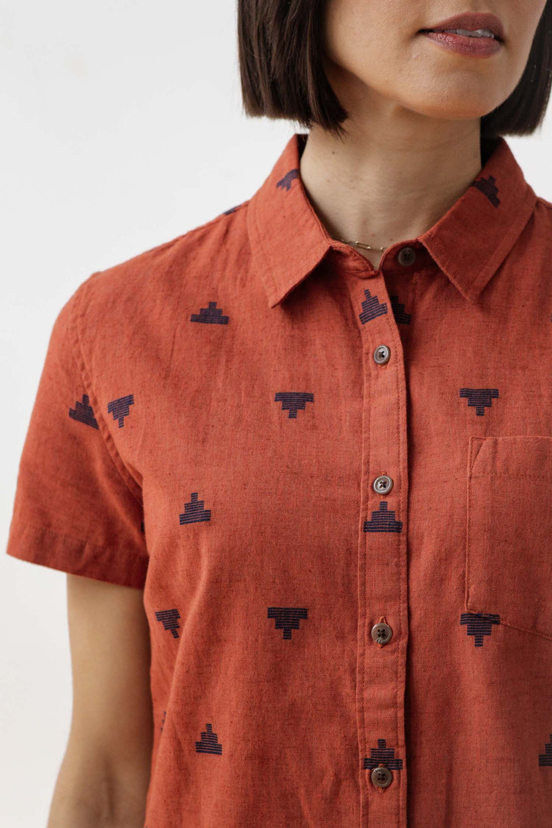 A woman wears the Bridge & Burn Lana Shirt in Rust Fil-Coupé Pyramids, a sustainable short-sleeve with pointed collar and dark geometric patterns, shown from shoulders to mouth.
