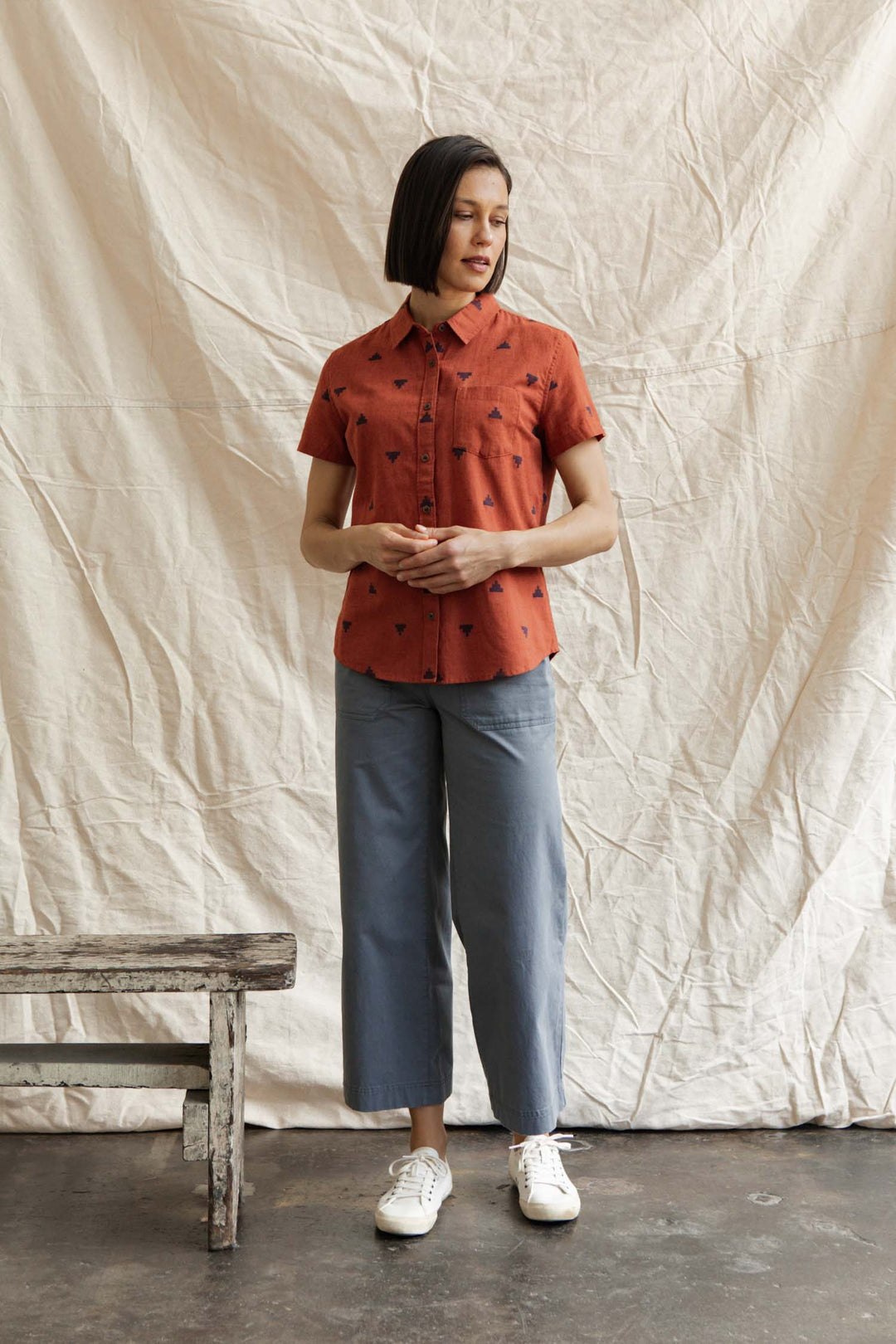 A woman stands indoors before a cream fabric backdrop, wearing the Bridge & Burn Lana Shirt in Rust Fil-Coupé Pyramids, light blue wide-leg pants, and white sneakers. A rustic wooden bench is to her left.