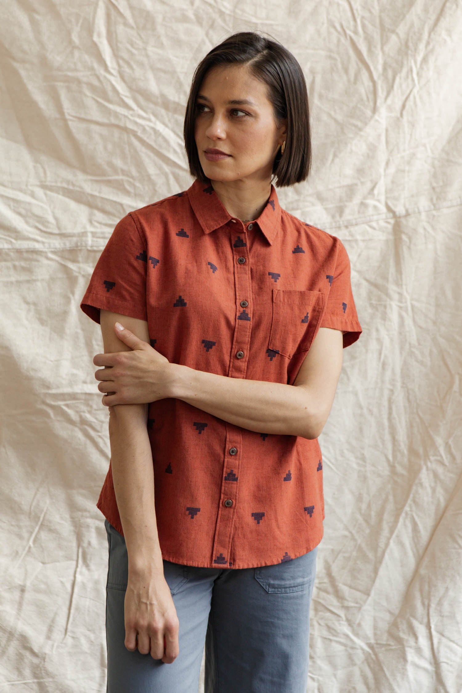A woman with short brown hair wears the Bridge & Burn Lana Shirt in Rust Fil-Coupé Pyramids, paired with light blue pants, standing before a beige textured fabric backdrop and gazing slightly to the side.
