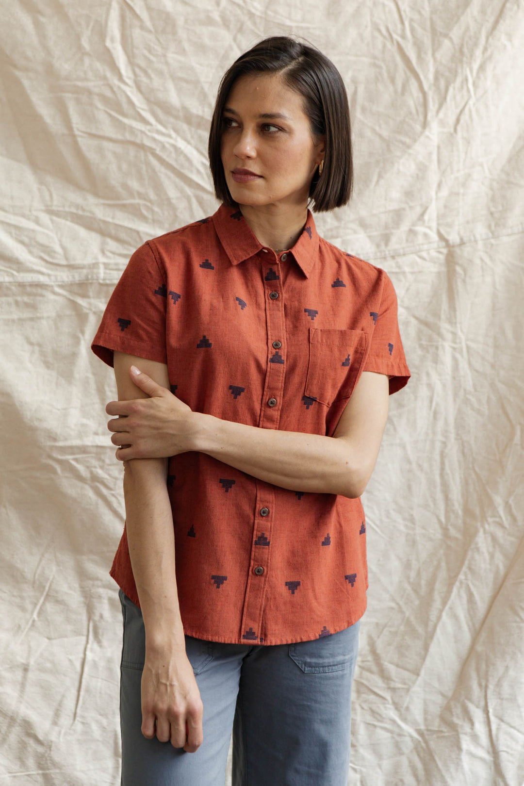 A woman with short brown hair wears the Bridge & Burn Lana Shirt in Rust Fil-Coupé Pyramids, paired with light blue pants, standing before a beige textured fabric backdrop and gazing slightly to the side.
