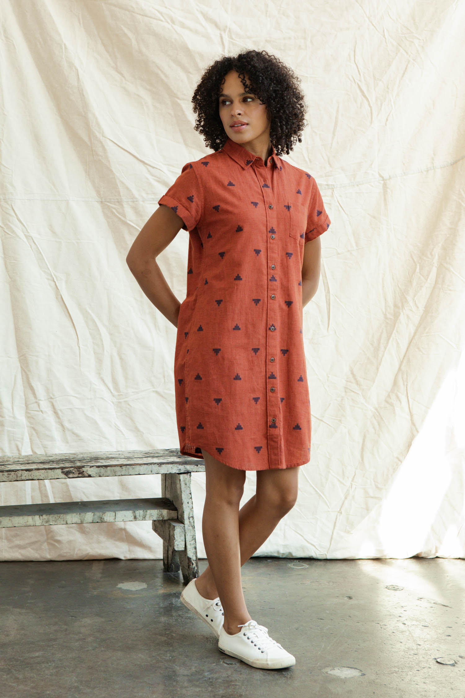 A woman with curly hair wears the Bridge & Burn Loren Shirt Dress in Rust Fil-Coupé Pyramids. Indoors, she stands before a cream backdrop by a rustic bench and pairs this sustainable button-front dress with white sneakers.