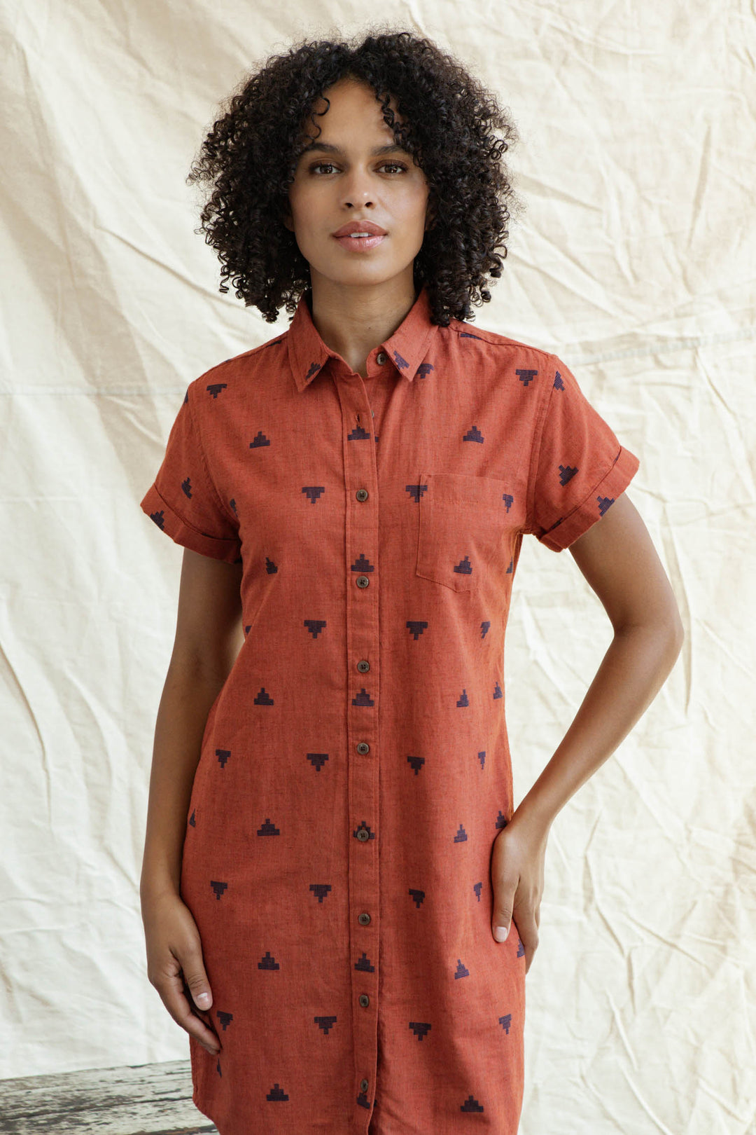 A woman with curly hair wears the Loren Shirt Dress in Rust Fil-Coupé Pyramids by Bridge & Burn, featuring short sleeves and a button front, standing before a cream-colored fabric backdrop.