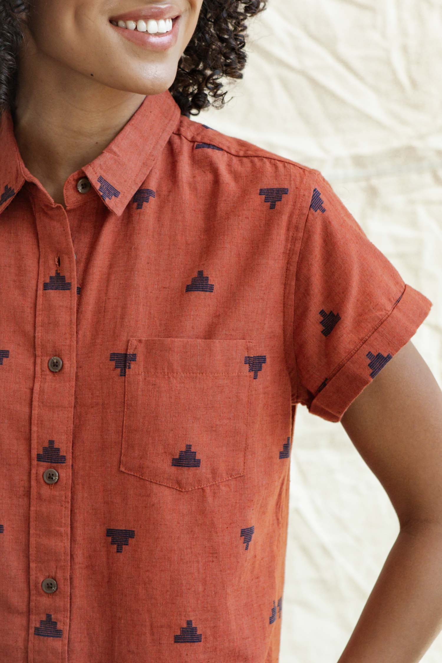 A person wears the Bridge & Burn Loren Shirt Dress in Rust Fil-Coupé Pyramids, a rust-toned, short-sleeved button-front dress with small navy geometric patterns, smiling against a light textured background. Upper body shown from shoulders to mid-torso.