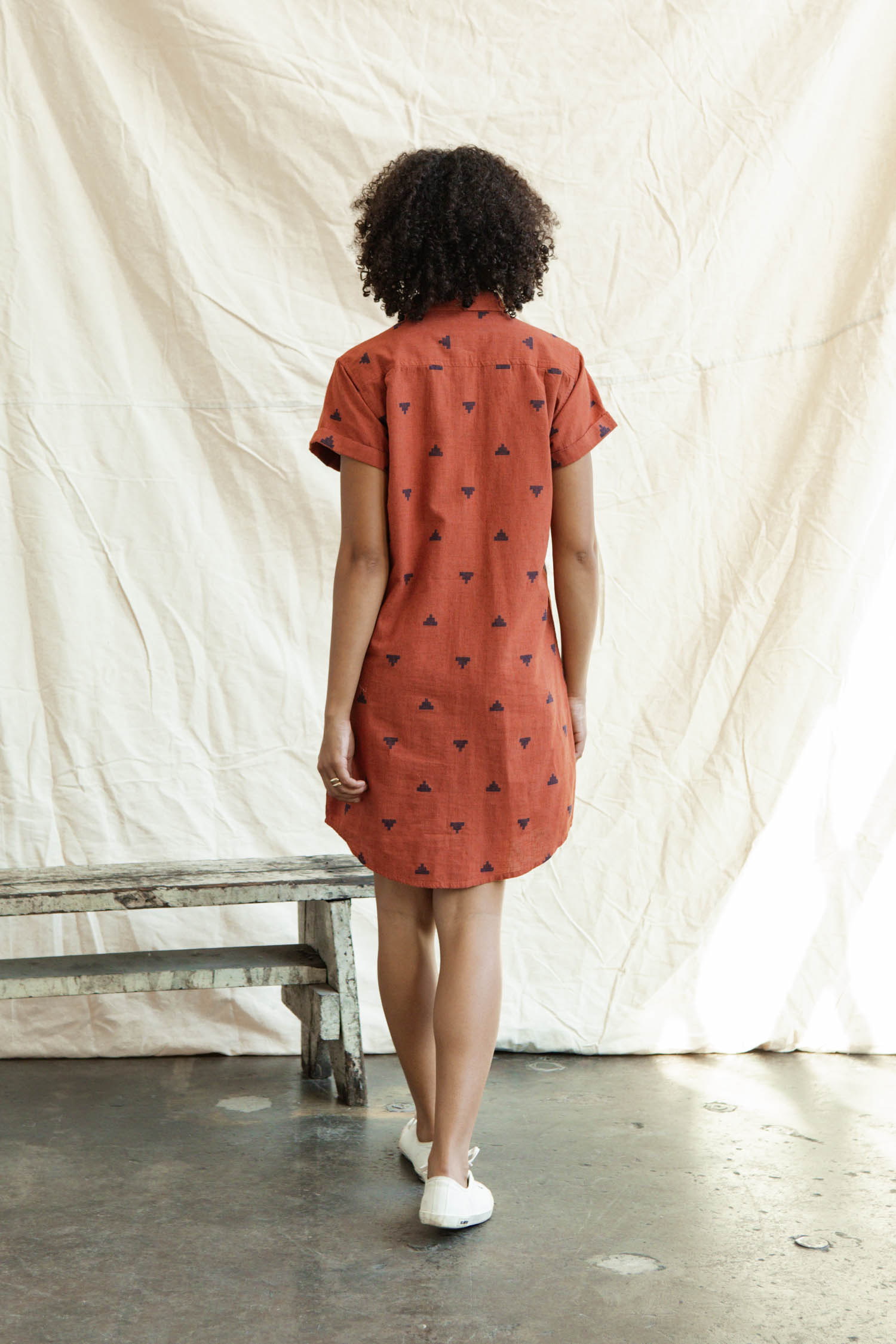 A person with curly hair stands facing away, wearing the Bridge & Burn Loren Shirt Dress in Rust Fil-Coupé Pyramids and white sneakers, indoors near a bench with a light fabric backdrop behind them.