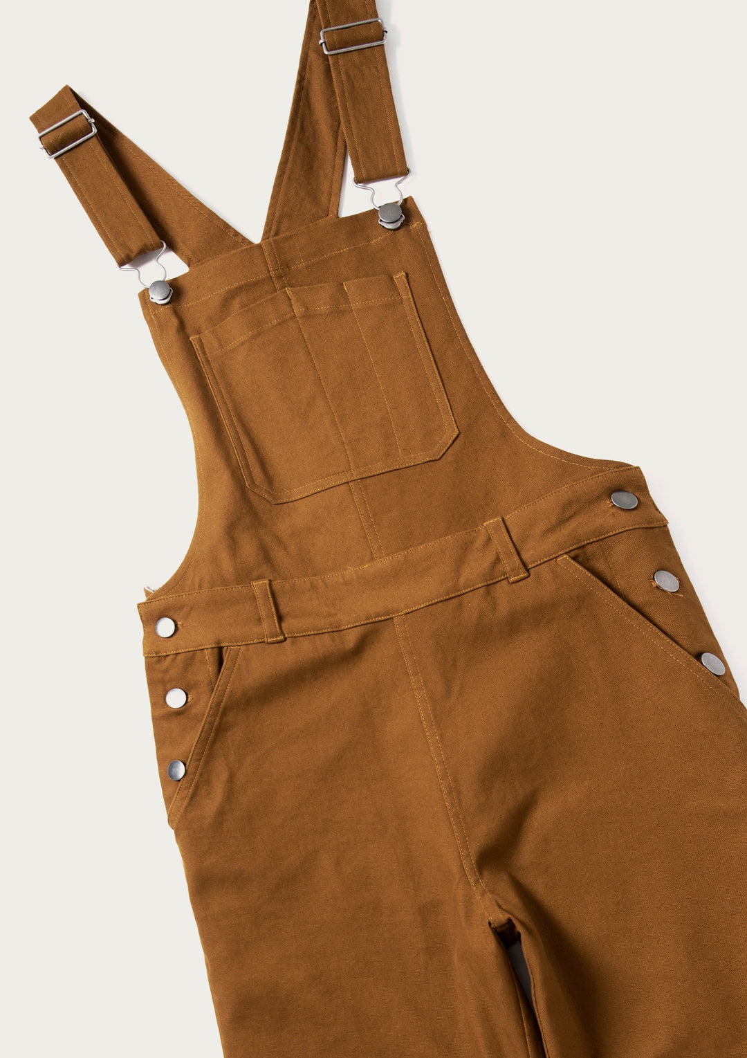 The Bridge & Burn Tobin Utility Overall in brown canvas features adjustable shoulder straps, a large front chest pocket, and metal hardware details, shown here laid flat against a neutral background.