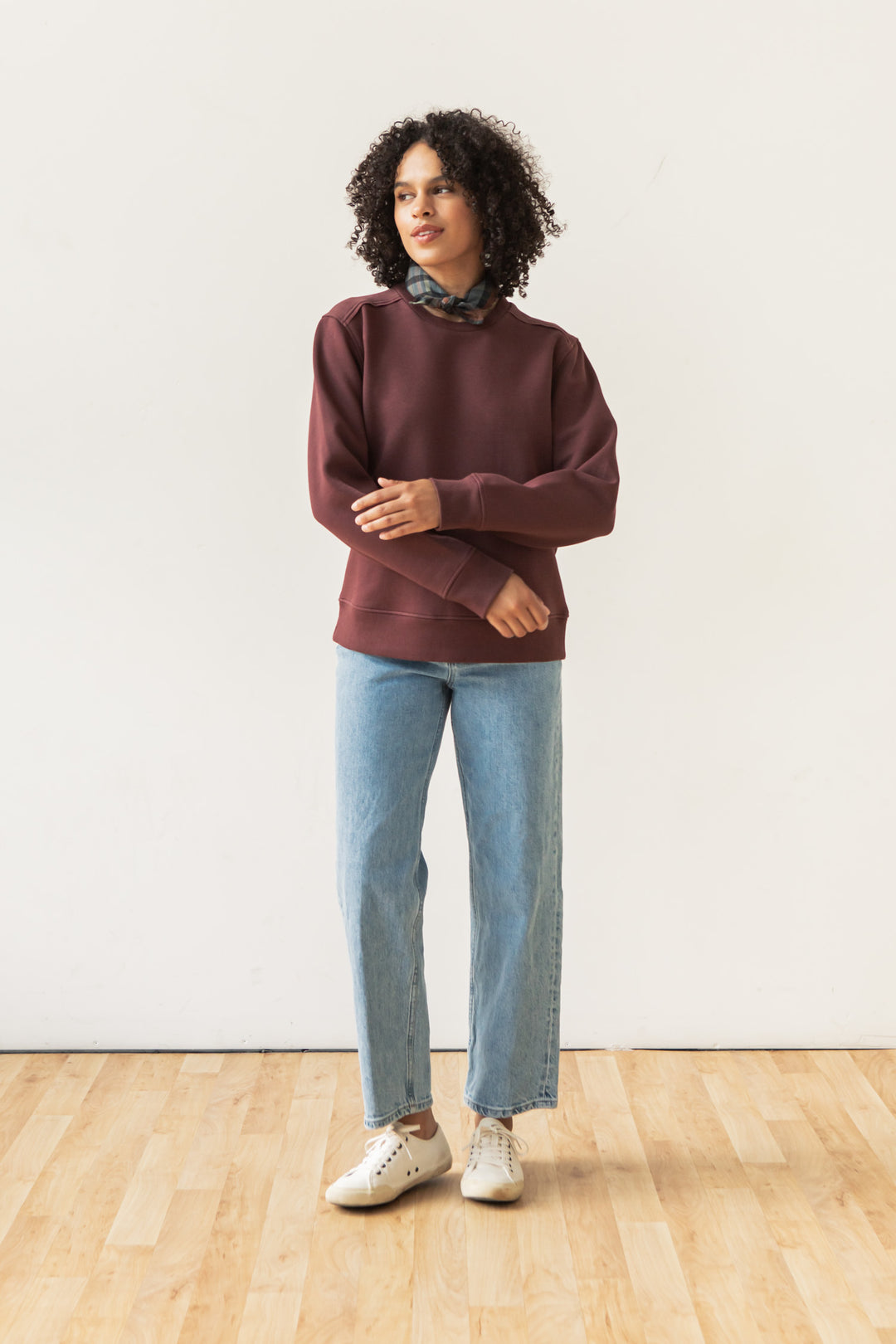 Wearing the Bailey Crew Sweatshirt in Burgundy by Bridge & Burn, light blue jeans, white sneakers, and a patterned scarf, a curly-haired person stands on a wooden floor against a plain white wall.