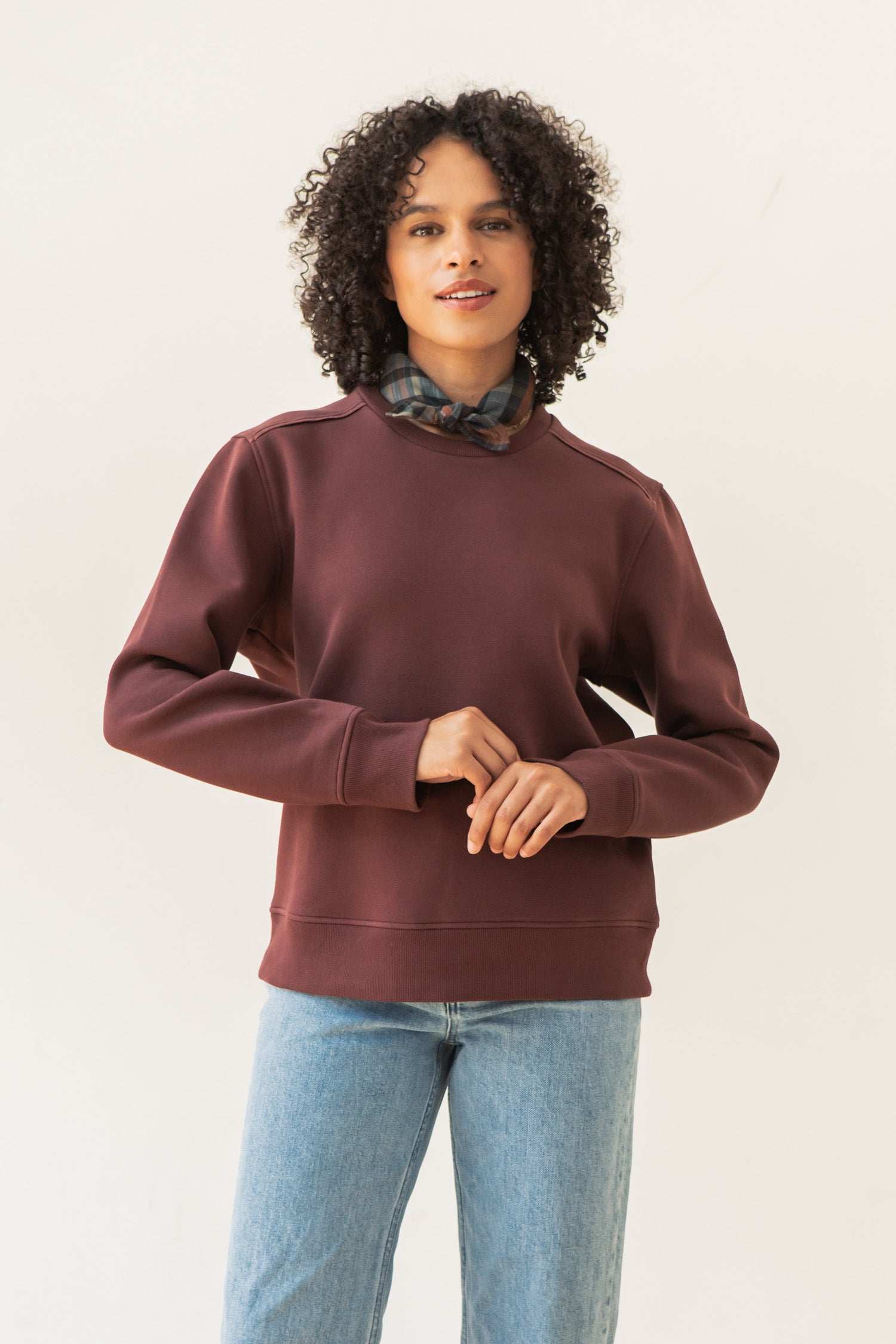 A person with curly hair wears a Bridge & Burn Bailey Crew Sweatshirt in Burgundy, light blue jeans, and a plaid scarf around their neck, standing against a plain white background.