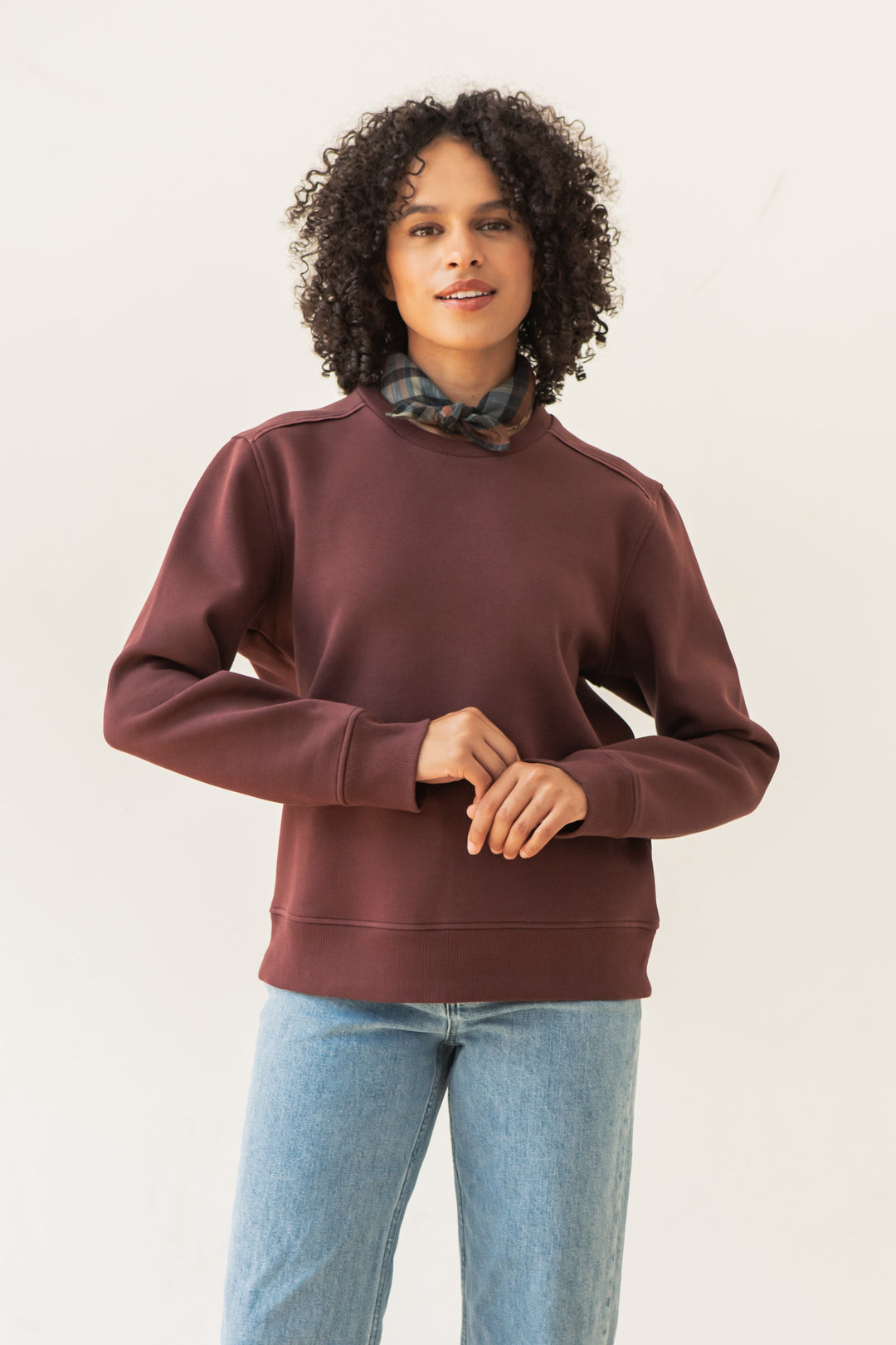 A person with curly hair wears a Bridge & Burn Bailey Crew Sweatshirt in Burgundy, light blue jeans, and a plaid scarf around their neck, standing against a plain white background.