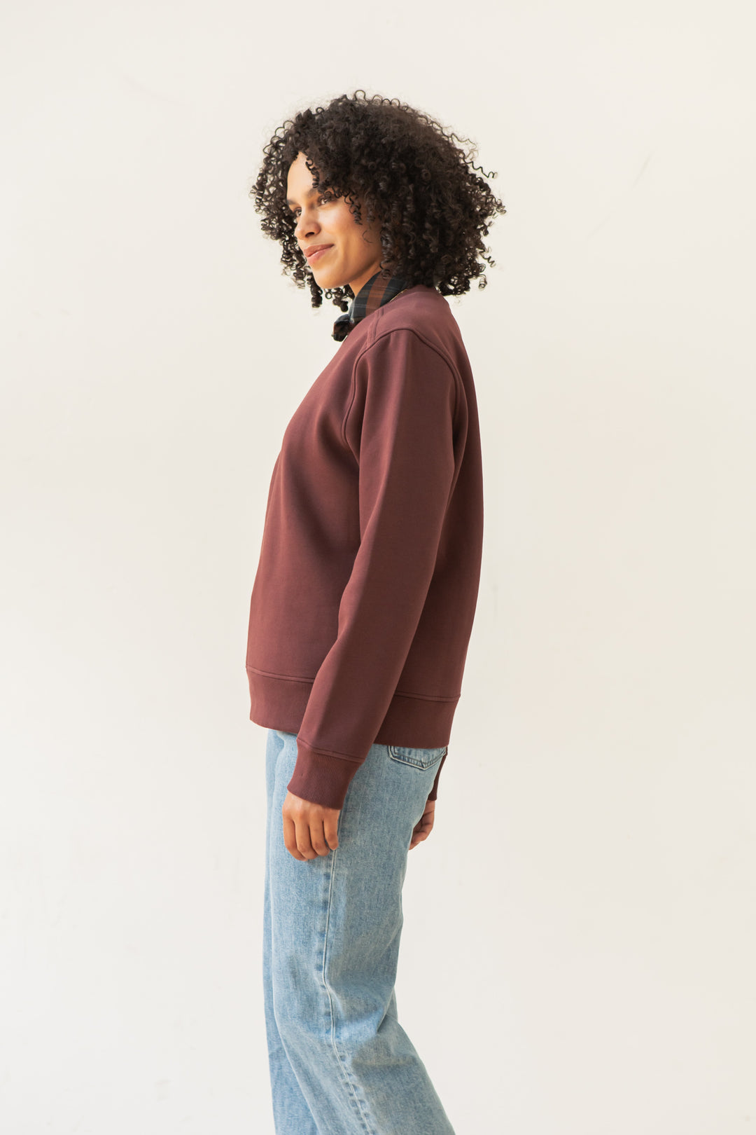 A person with curly hair stands sideways against a plain background, wearing the Bridge & Burn Bailey Crew Sweatshirt in burgundy and light blue jeans.