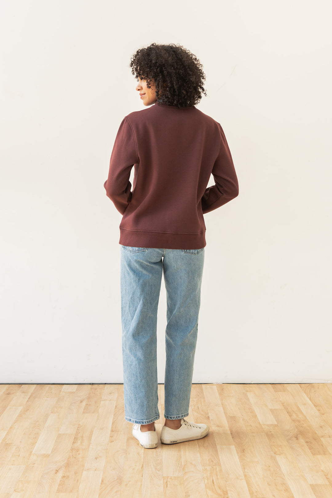A person with curly hair, wearing the Bridge & Burn Bailey Crew Sweatshirt in Burgundy, light blue jeans, and white sneakers, stands indoors on a wooden floor facing away from the camera against a plain white wall.