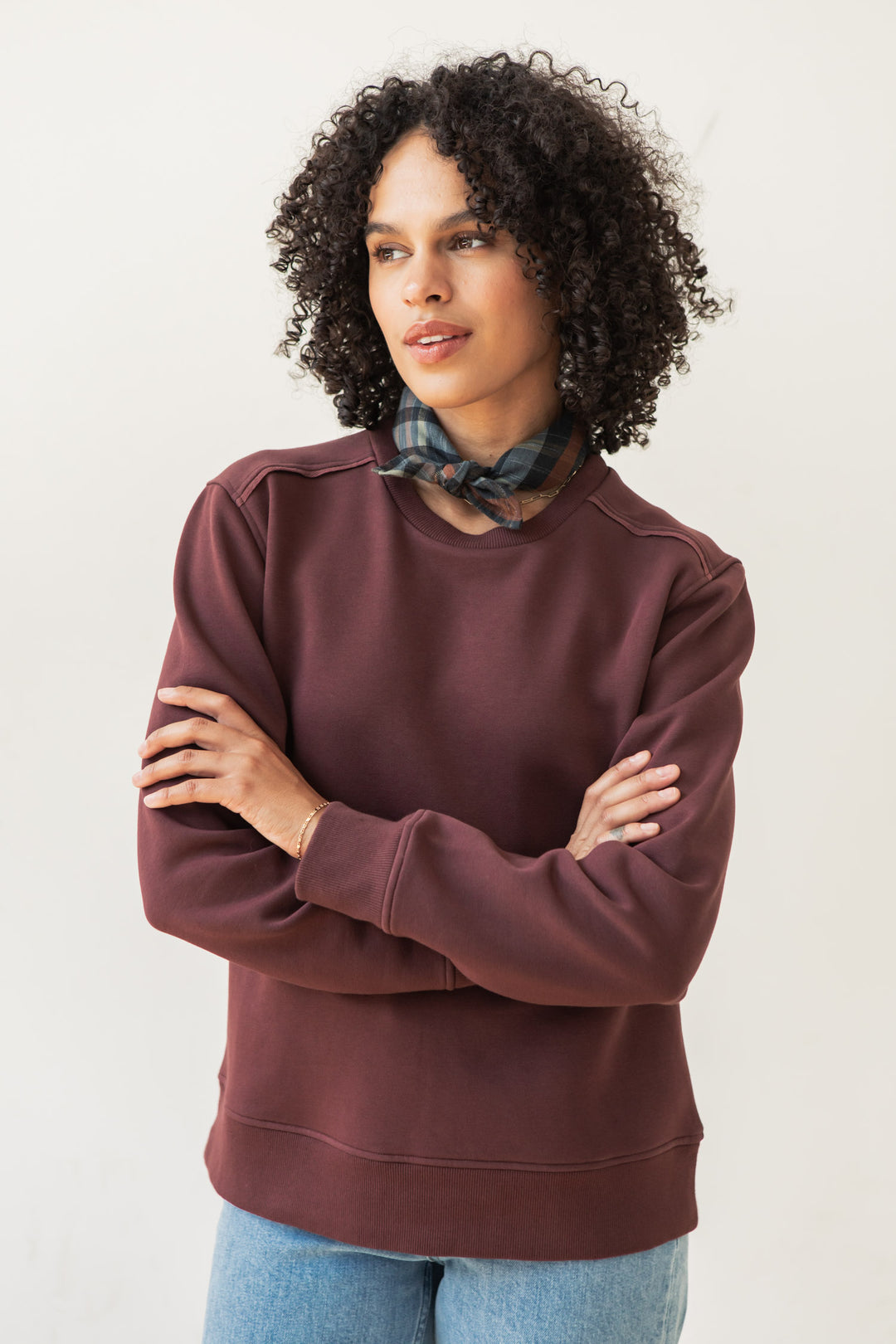 A woman with curly hair wears the Bridge & Burn Bailey Crew Sweatshirt in Burgundy, paired with blue jeans and a plaid scarf. She stands with arms crossed, looking to the side against a plain light background.
