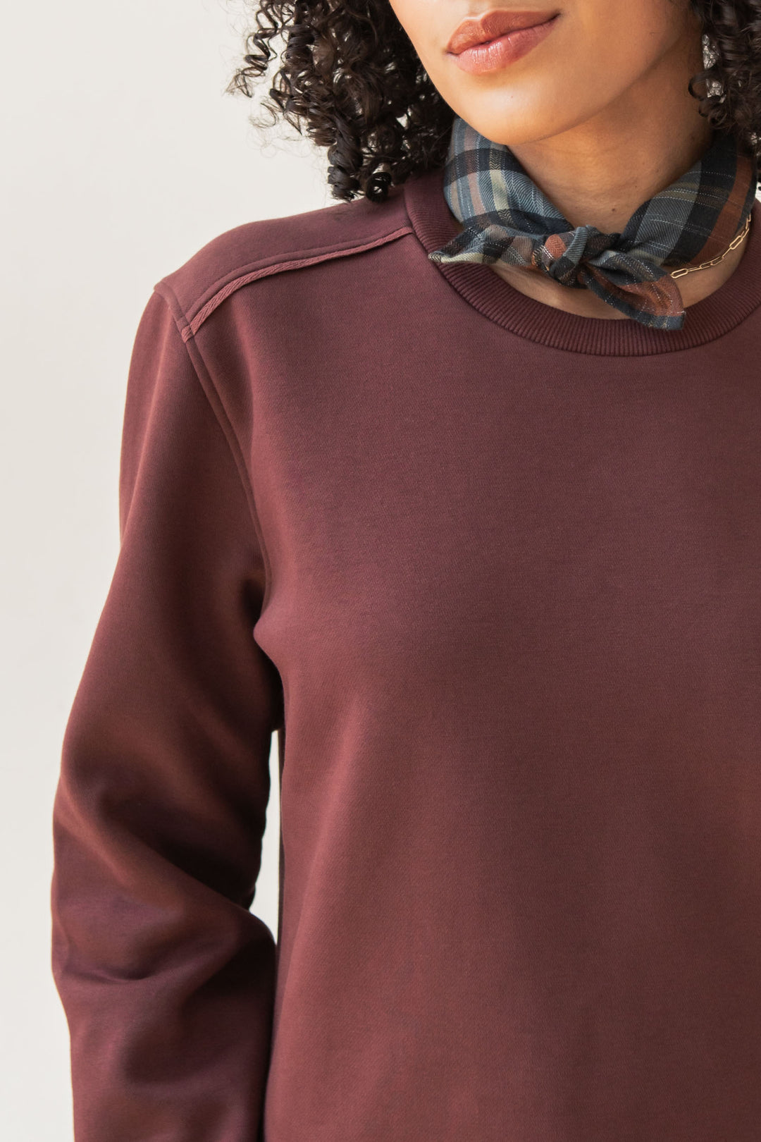 A woman with curly hair wears the Bridge & Burn Bailey Crew Sweatshirt in burgundy and a plaid neck scarf. Only her lower face, neck, and upper torso are visible against a plain light background.