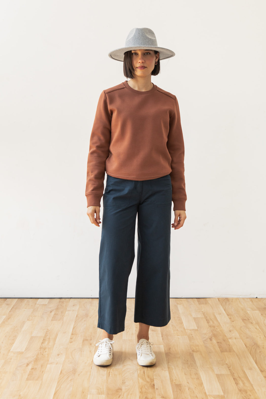 A woman stands indoors on a wooden floor, wearing Bridge & Burn's Bailey Crew Sweatshirt in Burnt Umber, a gray wide-brim hat, blue wide-leg pants, and white sneakers against a plain white wall.
