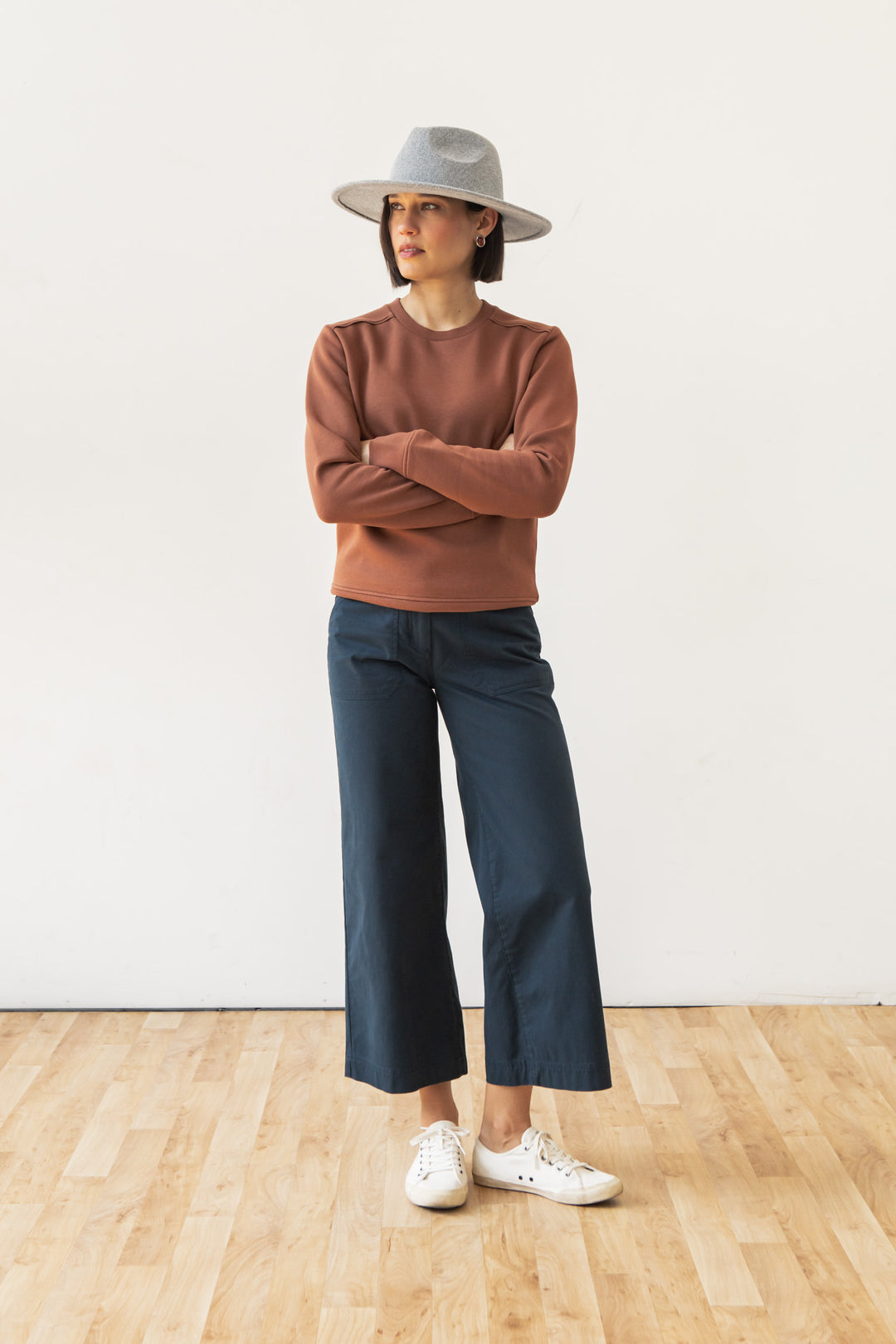 Wearing the Bridge & Burn Bailey Crew Sweatshirt in Burnt Umber, a person pairs it with a gray wide-brimmed hat, navy wide-leg pants, and white sneakers, standing on a wooden floor against a plain white background.