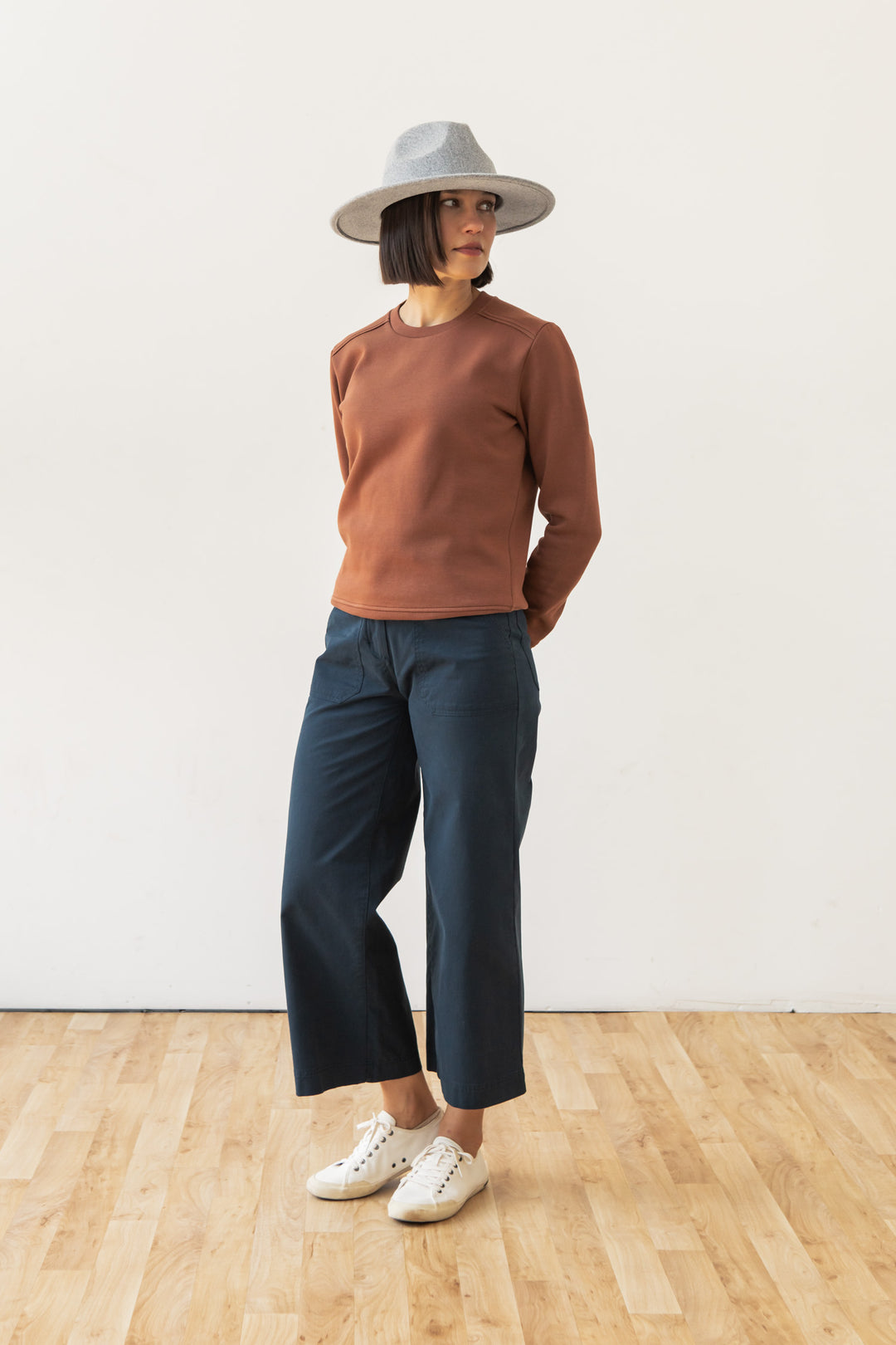 A woman wears the Bridge & Burn Bailey Crew Sweatshirt in Burnt Umber, styled with dark blue cropped pants and white sneakers, standing on a wooden floor against a plain white wall and looking to the side.