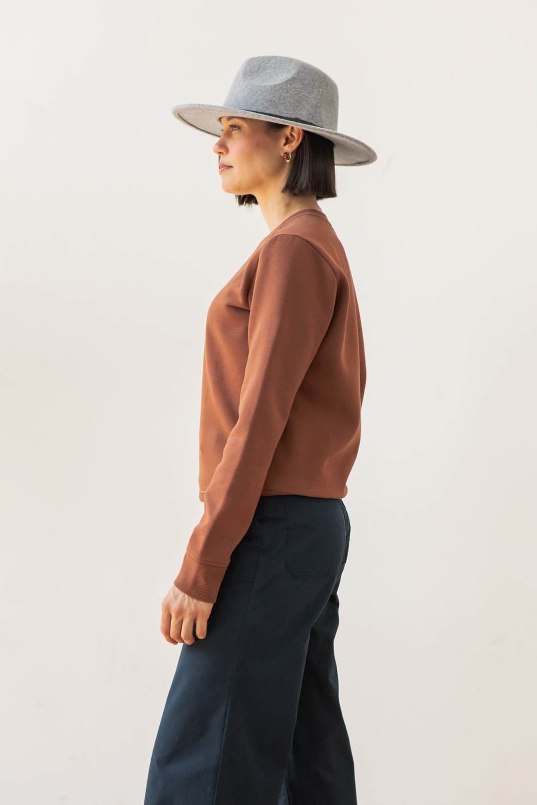 A person with short dark hair wears a gray wide-brimmed hat and the Bridge & Burn Bailey Crew Sweatshirt in Burnt Umber, paired with dark pants, while standing in profile against a plain light background.