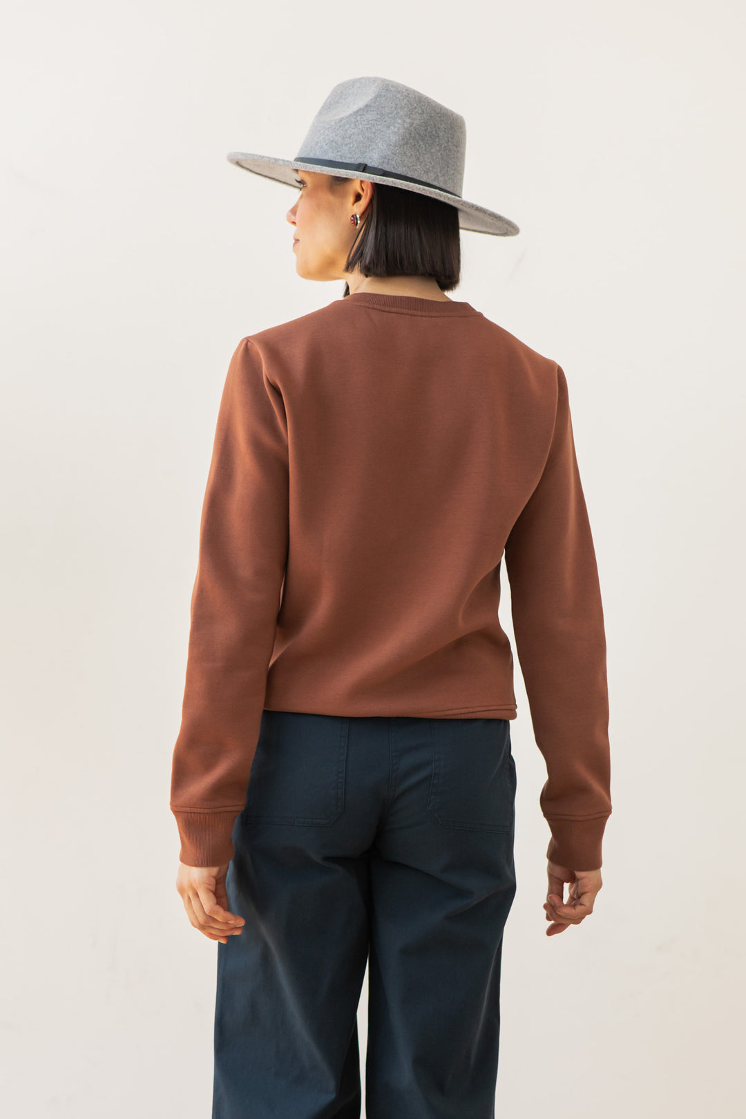 A person wearing the Bridge & Burn Bailey Crew Sweatshirt in Burnt Umber, a gray wide-brimmed hat, and dark pants stands facing away from the camera against a plain light background.
