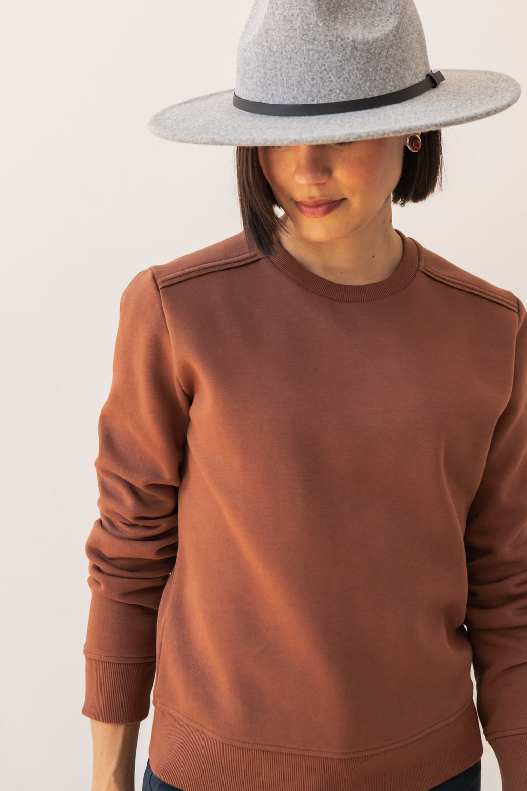 A woman in the Bridge & Burn Bailey Crew Sweatshirt in Burnt Umber and a grey wide-brimmed hat looks down, partially covering her face with the brim against a plain light background.