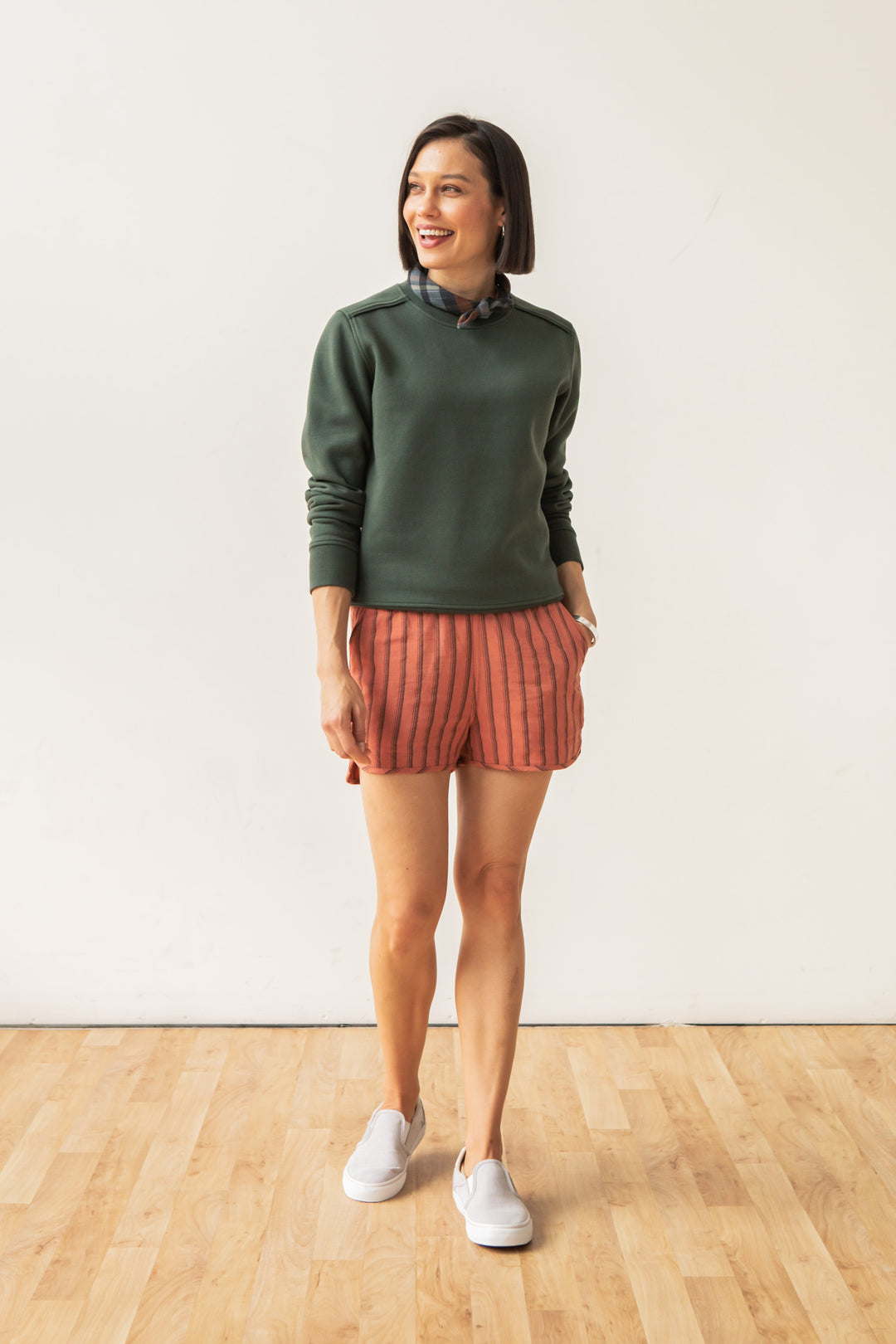 A woman stands on a wooden floor against a white wall, wearing the Bridge & Burn Bailey Crew Sweatshirt in Forest Night, pink striped shorts, and white slip-on shoes. She smiles and looks to the side with one hand in her pocket.