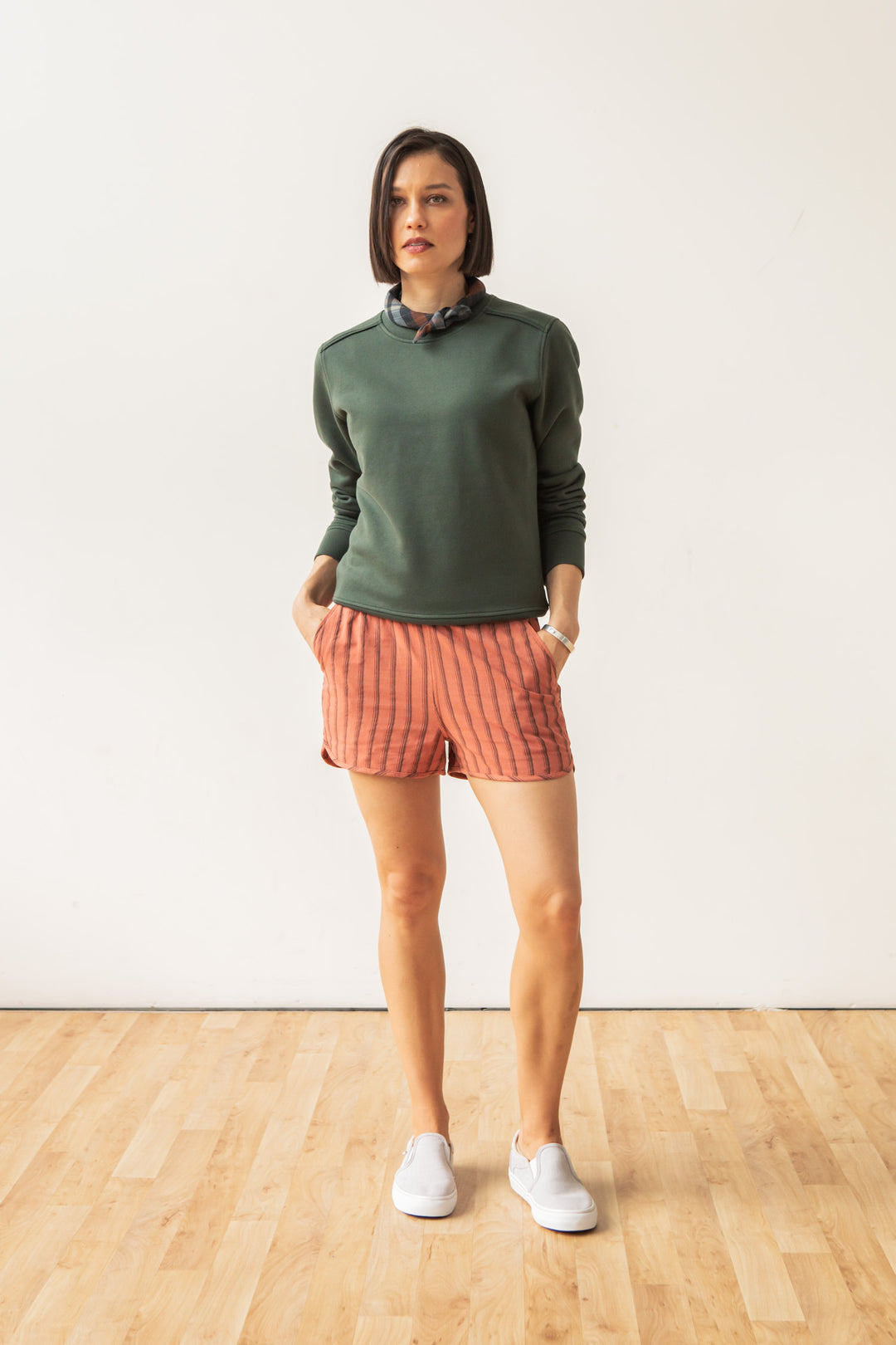 A woman stands indoors on a wooden floor wearing the Bridge & Burn Bailey Crew Sweatshirt in Forest Night, paired with pink striped shorts and white sneakers. Hands in her shorts pockets, she gazes to the side with a neutral expression.