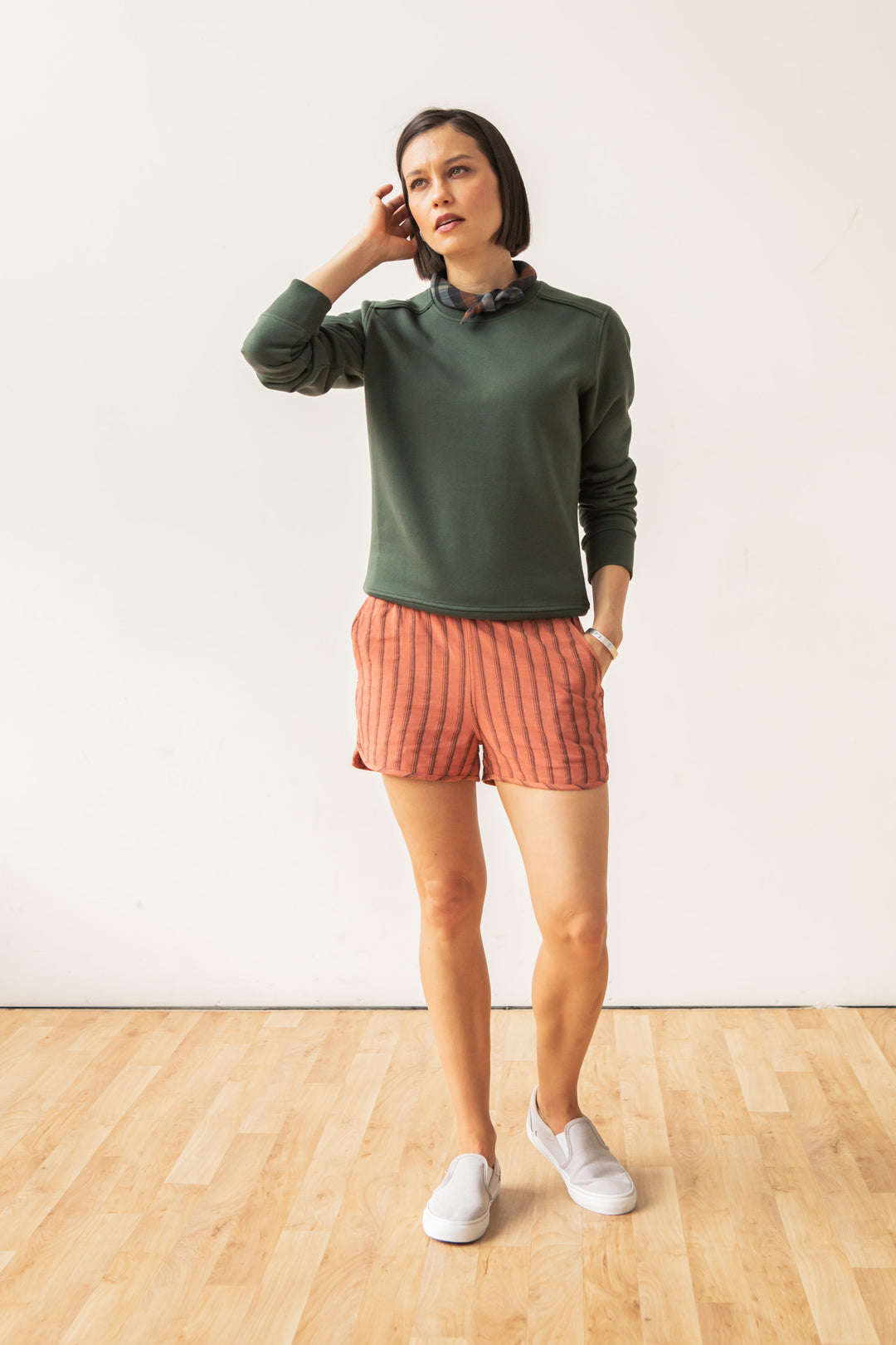 A woman stands indoors on a wooden floor, wearing the Bailey Crew Sweatshirt in Forest Night by Bridge & Burn, coral striped shorts, white slip-on shoes, and a dark scrunchie. One hand touches her head as she maintains a neutral expression.