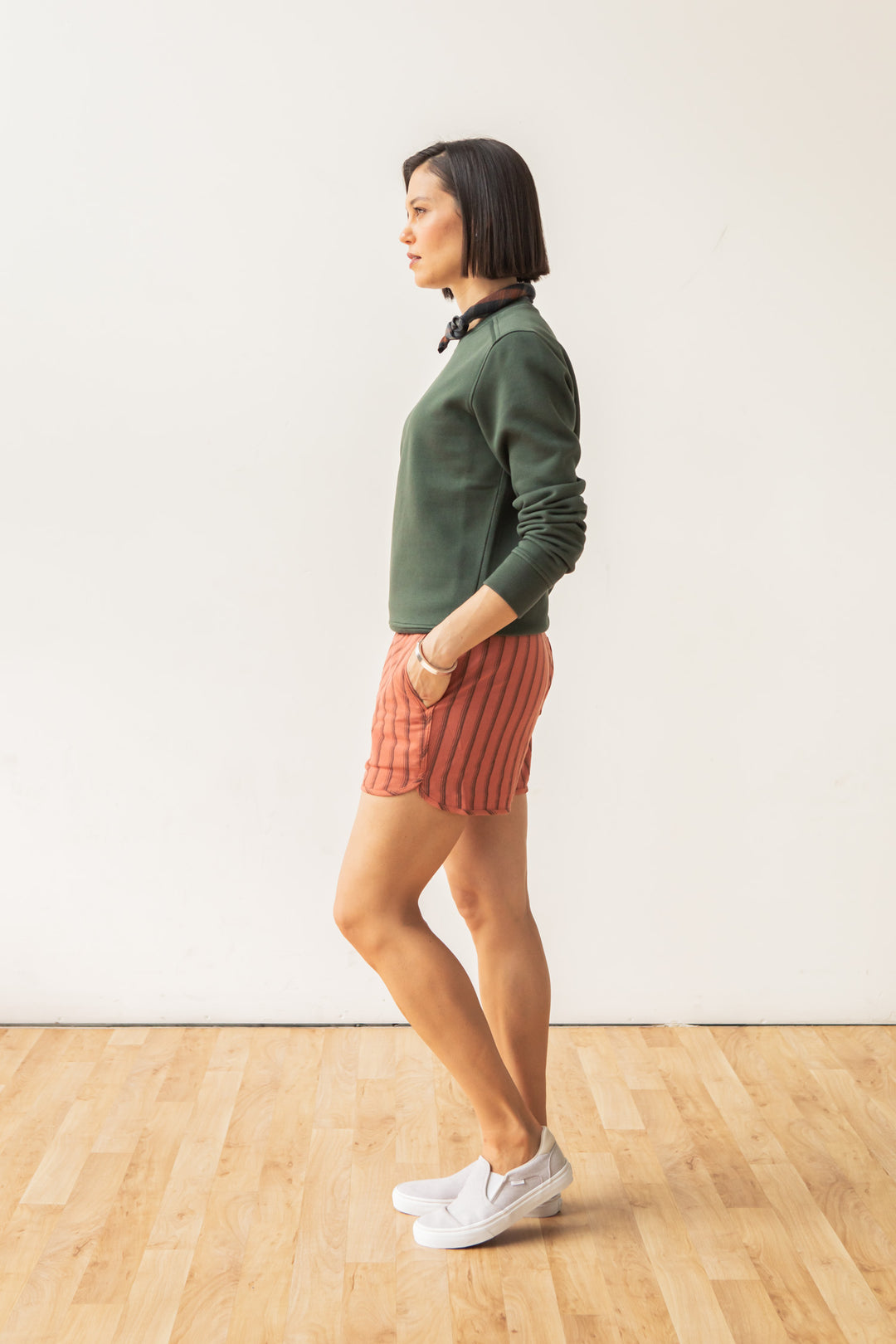 A woman with short dark hair stands in profile against a white wall, wearing the Bridge & Burn Bailey Crew Sweatshirt in Forest Night, red striped shorts, and white sneakers, hands in pockets on a wooden floor.
