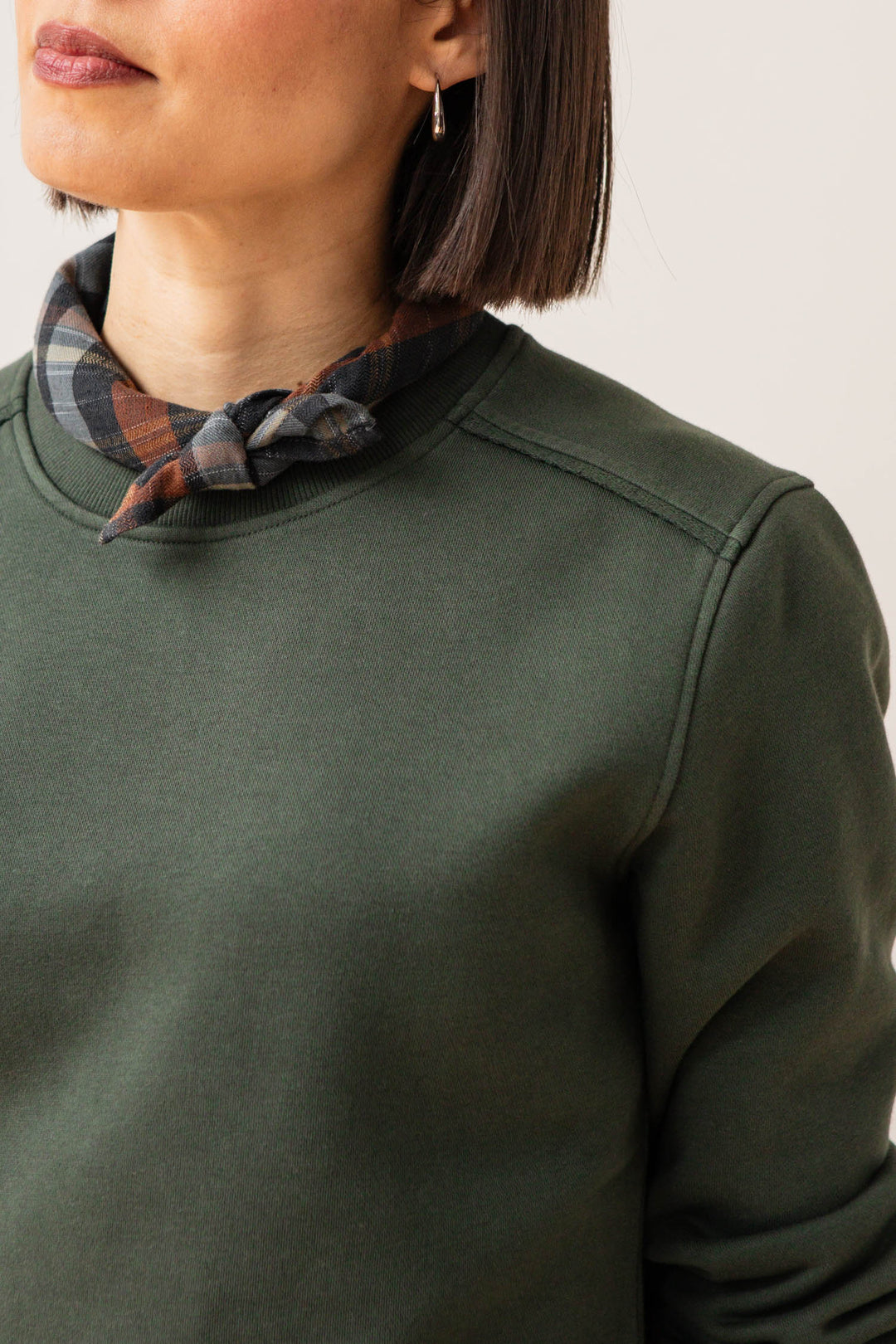 A woman with straight, chin-length dark hair wears the Bridge & Burn Bailey Crew Sweatshirt in Forest Night and a plaid scarf tied at her neck. Only the lower half of her face and upper torso are visible.