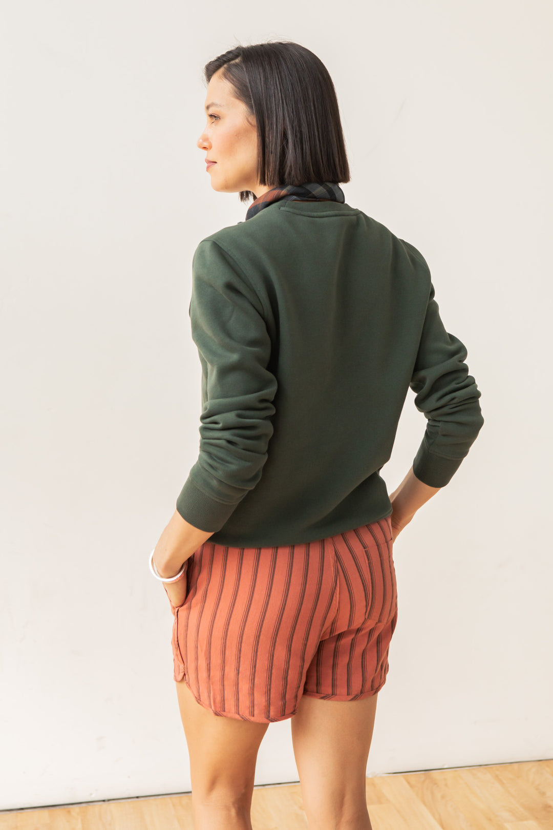 A person with a straight bob stands sideways, wearing the Bridge & Burn Bailey Crew Sweatshirt in Forest Night and rust-striped shorts, hands in pockets, against a plain light background.