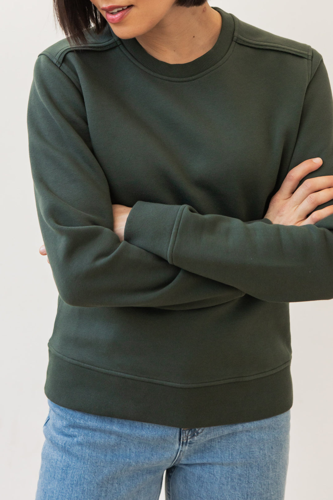 A person with short dark hair, wearing the Bridge & Burn Bailey Crew Sweatshirt in Forest Night and light blue jeans, stands arms crossed against a plain light background. Their face is partially out of the frame.