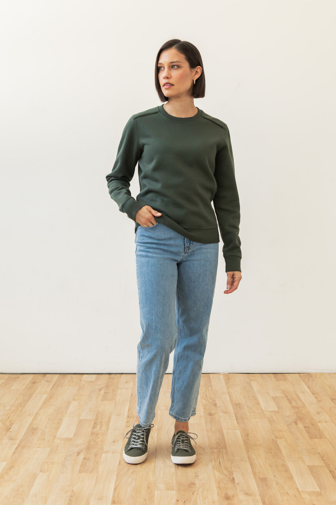 A woman with short dark hair wears the Bridge & Burn Bailey Crew Sweatshirt in Forest Night, paired with light blue jeans and green sneakers. She stands indoors on a wooden floor against a white wall, one hand in her pocket, looking to the side.