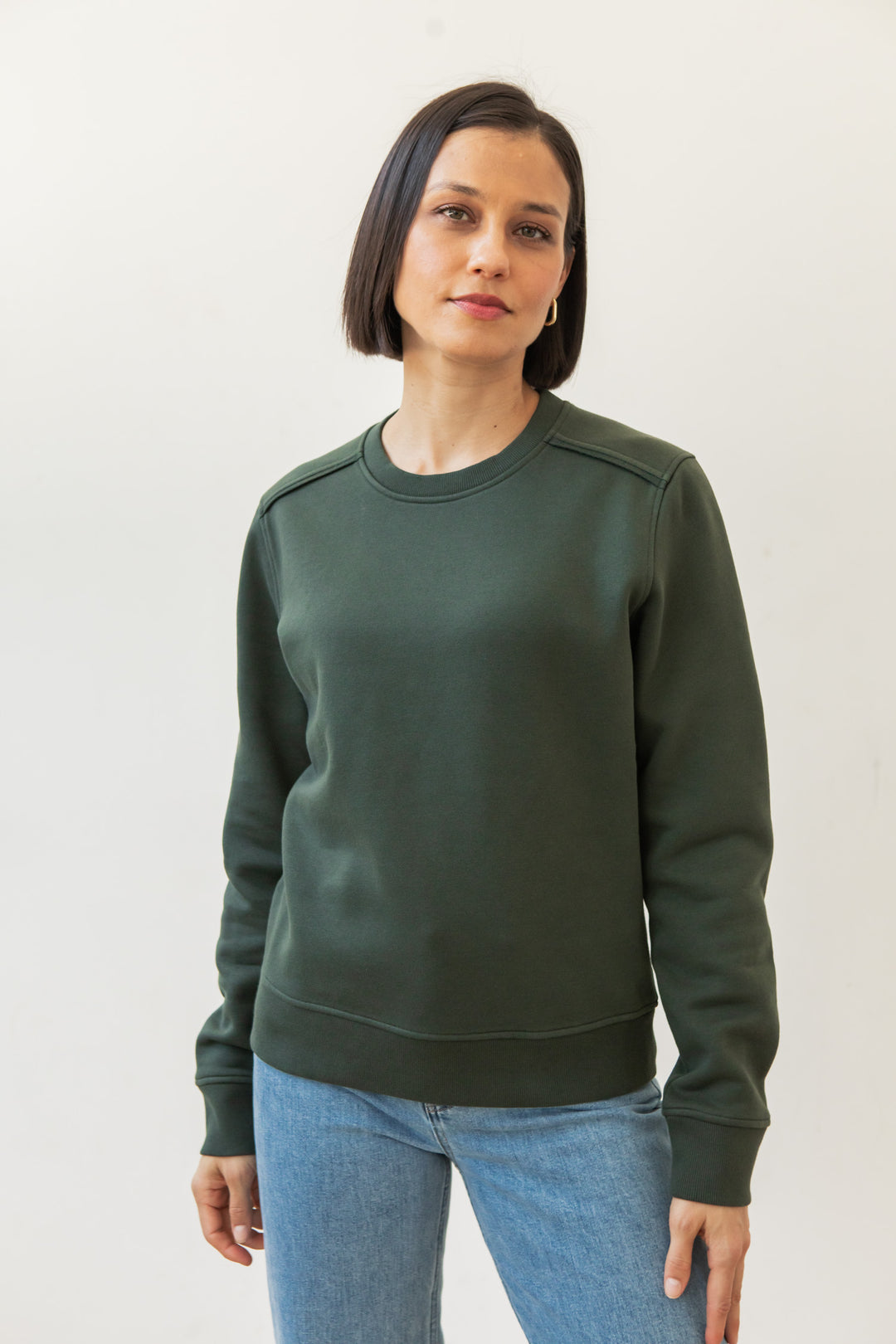 A woman with straight dark hair wears the Bridge & Burn Bailey Crew Sweatshirt in Forest Night and blue jeans, standing against a plain white background and looking at the camera with a neutral expression.