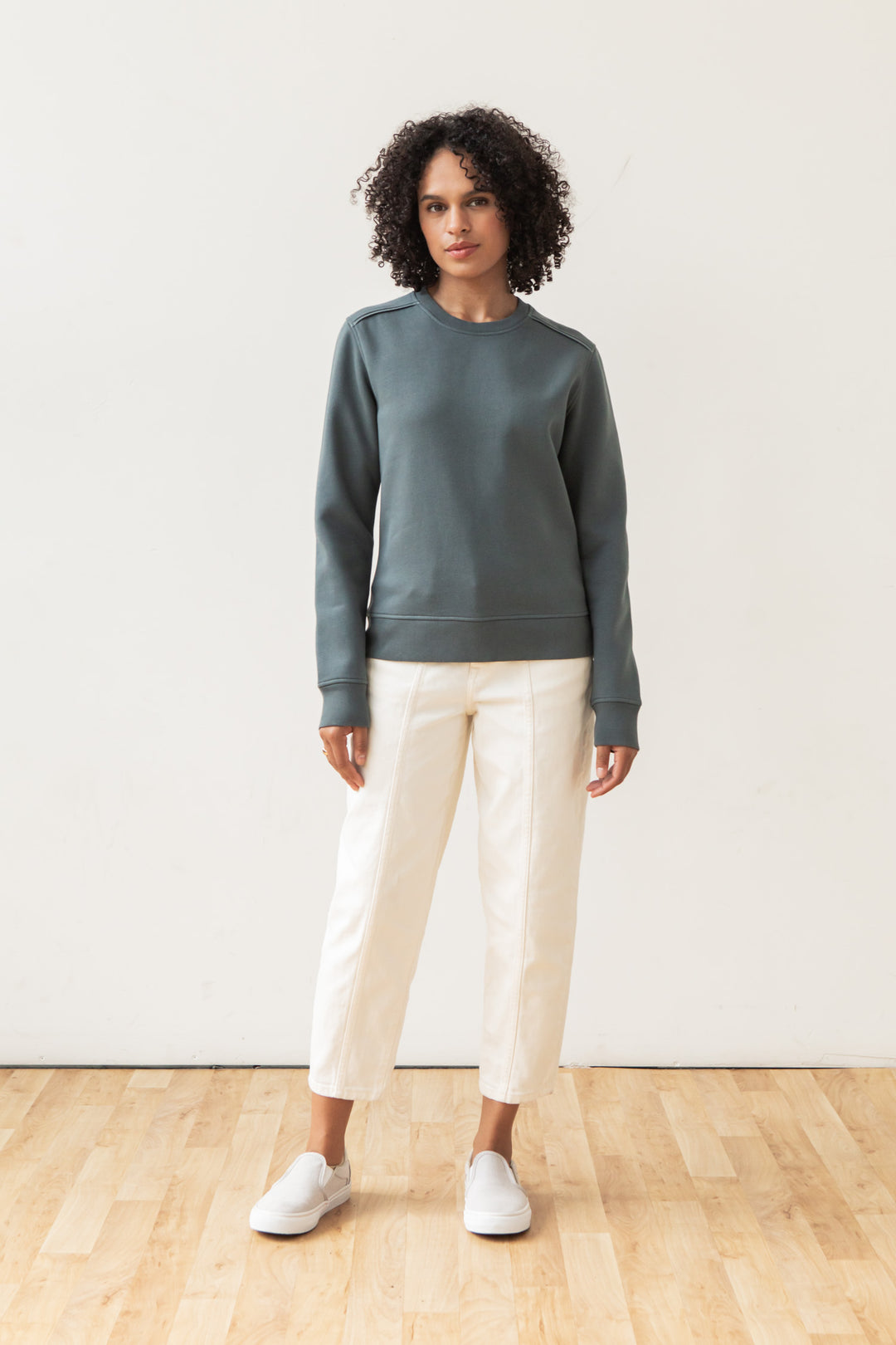 A person with curly hair stands indoors on a wooden floor, wearing the Bridge & Burn Bailey Crew Sweatshirt in Slate, white pants, and white slip-on shoes against a plain white wall.
