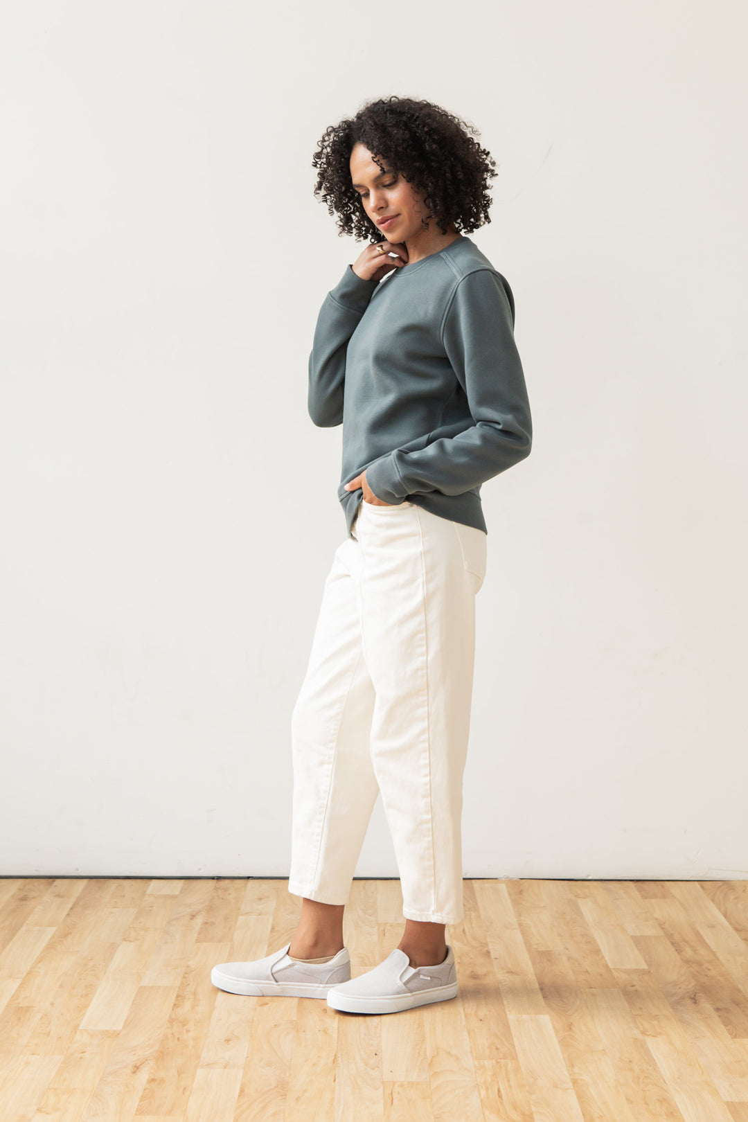 A woman with curly hair stands indoors on a wooden floor, wearing the Bridge & Burn Bailey Crew Sweatshirt in Slate, white pants, and gray slip-on shoes. She looks down thoughtfully with one hand near her chin and the other in her pocket.