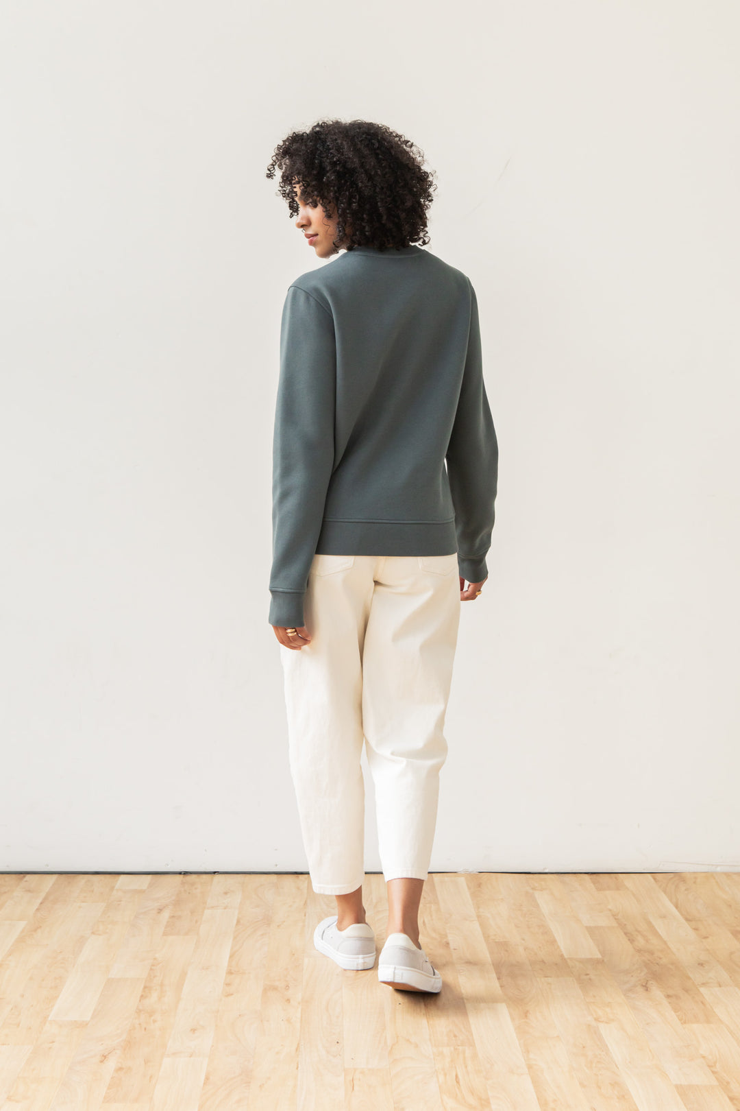 A person with curly hair, dressed in the Bridge & Burn Bailey Crew Sweatshirt in Slate, white pants, and gray slip-on shoes, stands on a wooden floor facing away from the camera against a plain white wall.