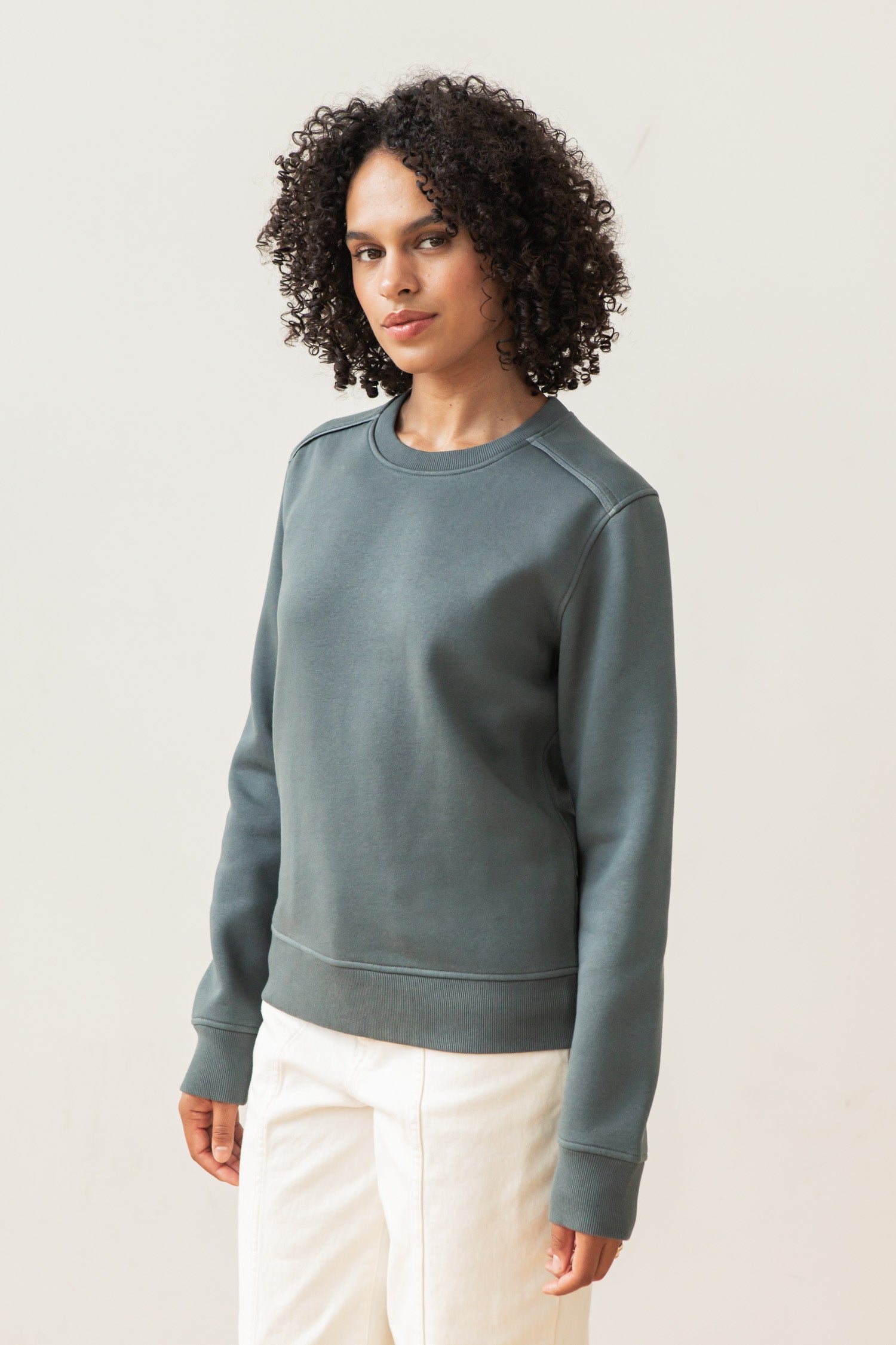 A person with curly hair wears the Bridge & Burn Bailey Crew Sweatshirt in Slate and off-white pants, standing against a plain light background and looking at the camera with a neutral expression.