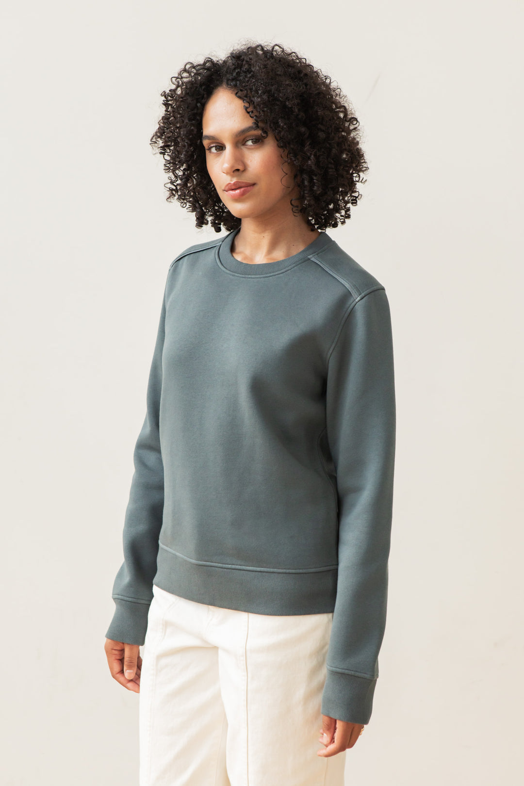 A person with curly hair wears the Bridge & Burn Bailey Crew Sweatshirt in Slate and off-white pants, standing against a plain light background and looking at the camera with a neutral expression.