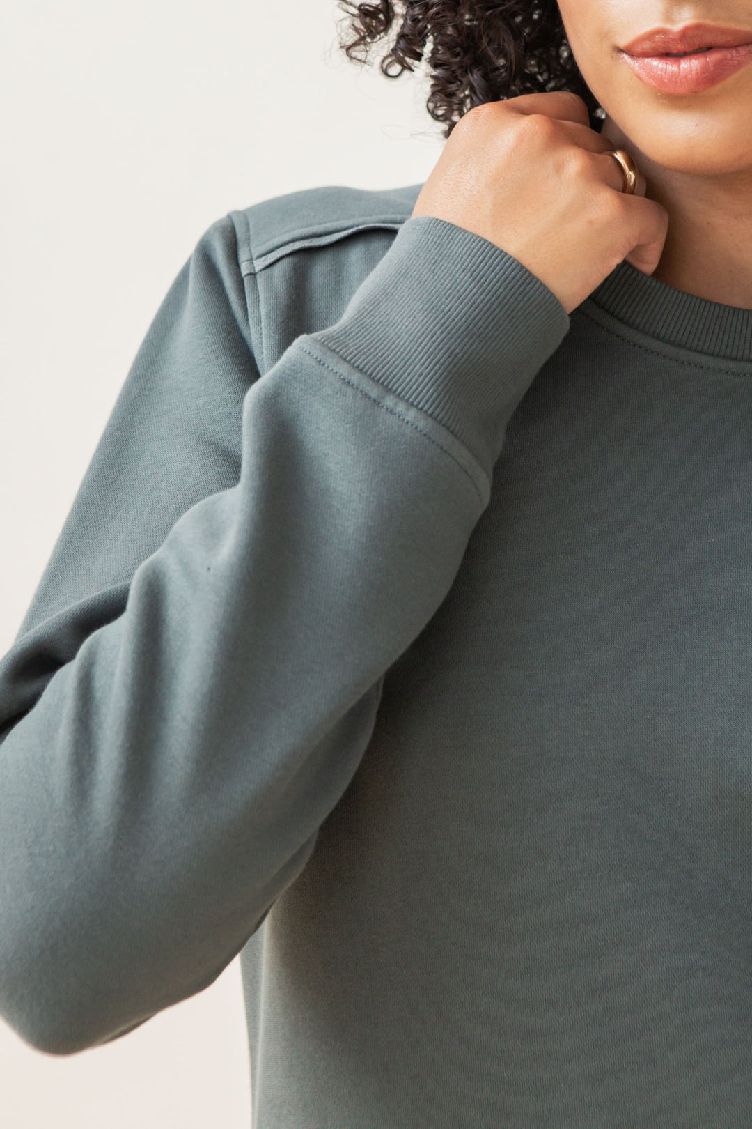 A person with curly hair gently holds the neckline of a Bridge & Burn Bailey Crew Sweatshirt in Slate, highlighting its textured organic cotton fabric and stitching against a plain, neutral background.