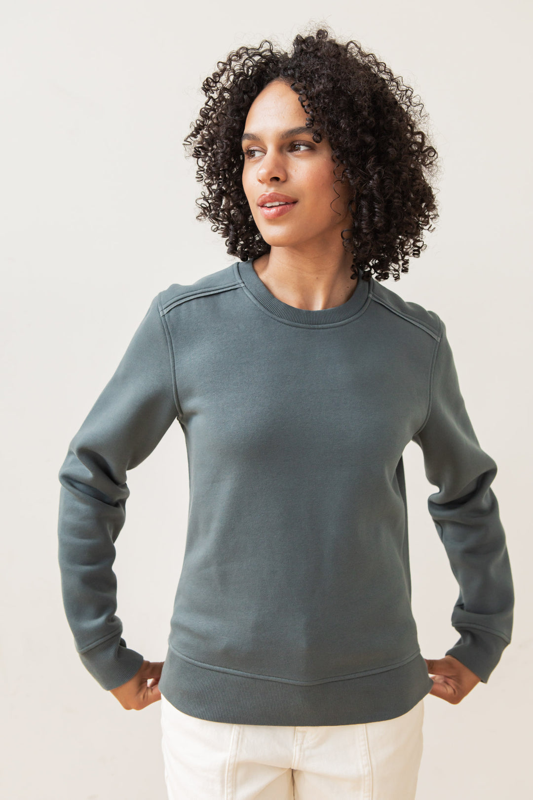 A person with curly hair wears the Bridge & Burn Bailey Crew Sweatshirt in Slate and light pants, standing against a plain light background, looking slightly to the side with hands on their hips.