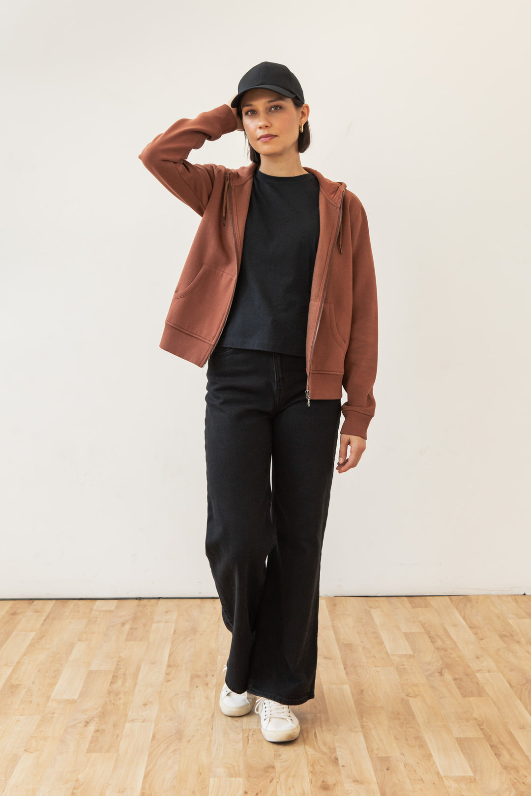 A person wearing the Bridge & Burn Mason Hoodie in Burnt Umber, a black cap, black t-shirt, black wide-leg pants, and white sneakers stands on a wooden floor against a white wall, with one hand touching their head.