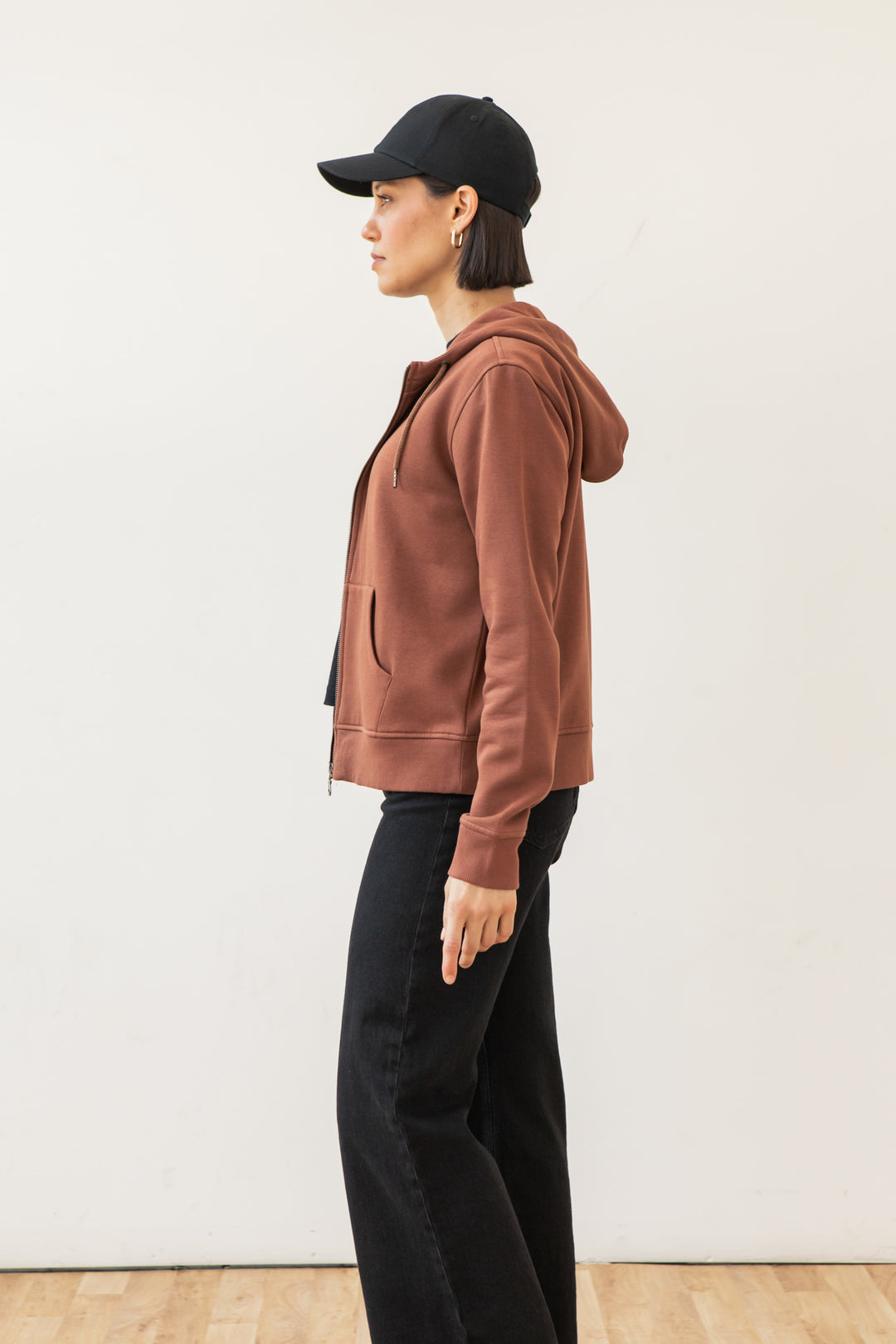 A person in the Bridge & Burn Mason Hoodie in Burnt Umber, black pants, and a black cap stands in profile against a plain white wall on a wooden floor.