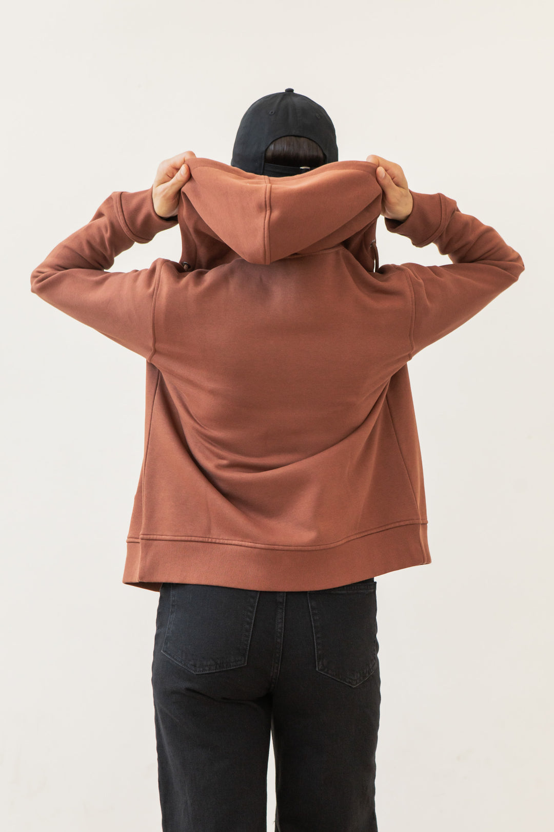 A person wearing the Bridge & Burn Mason Hoodie in Burnt Umber and black jeans stands facing away, lifting the hood over their head. They also wear a black cap. The background is plain white.
