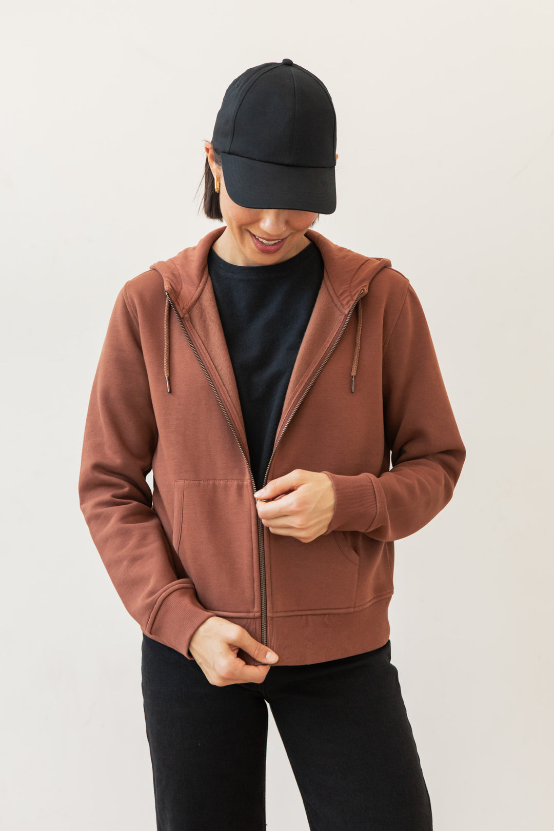 A person wearing a black baseball cap, black T-shirt, and Bridge & Burn's Mason Hoodie in Burnt Umber stands against a plain light background, looking down and adjusting the hoodie zipper.